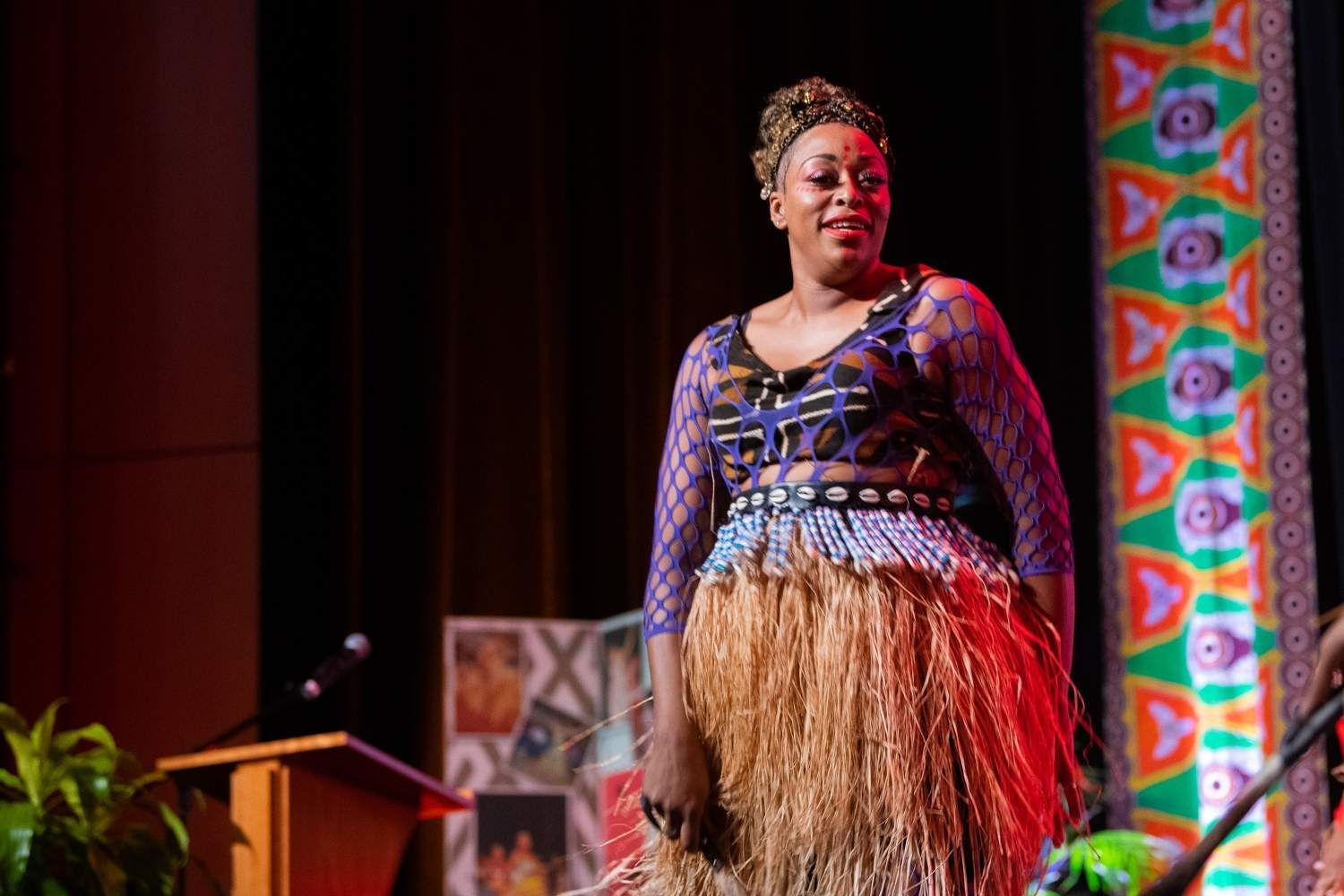 Woman in cultural attire on stage, smiling, speaking into a microphone. Decorated backdrop.