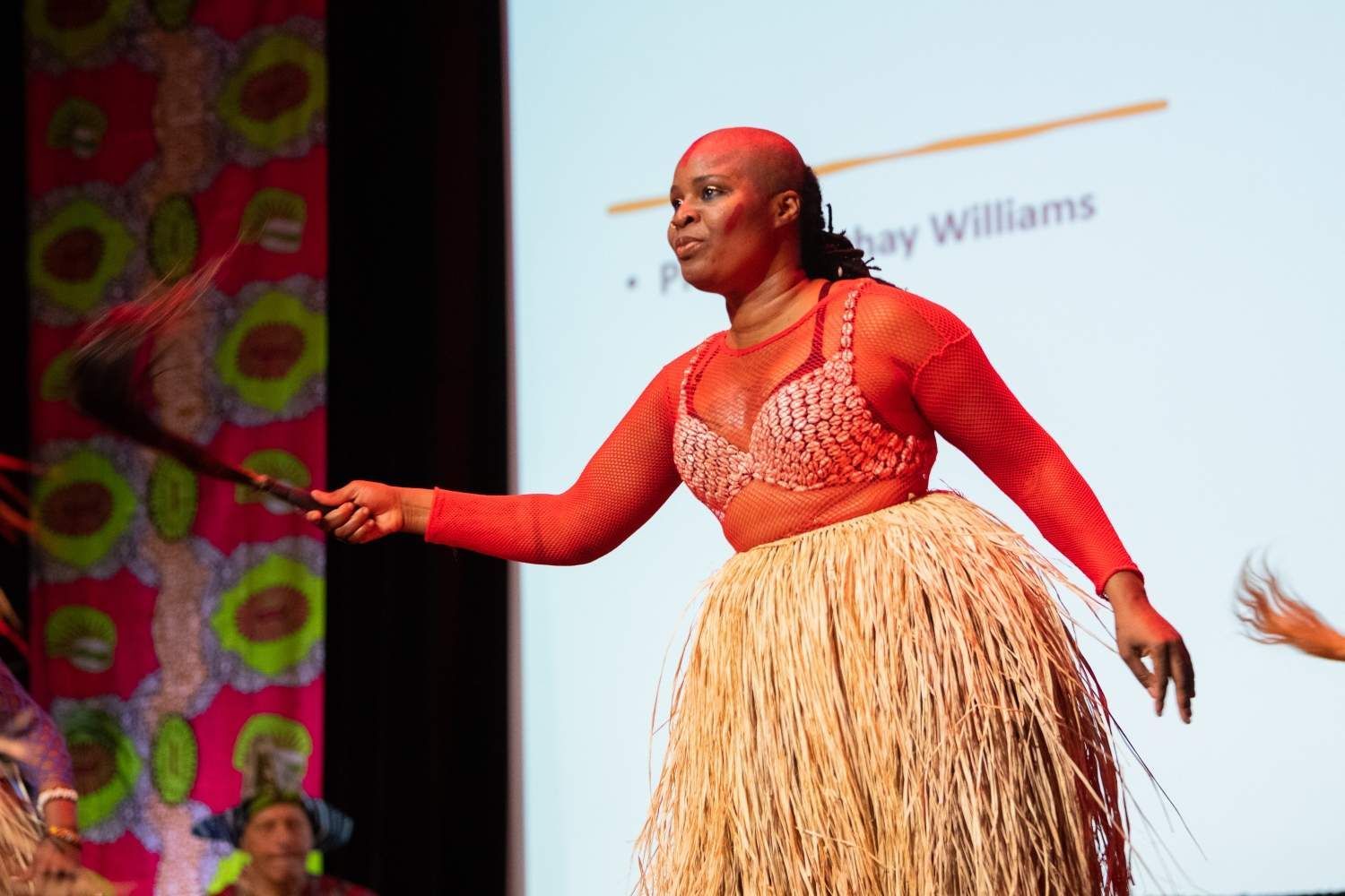Woman in red top and straw skirt dances on stage, holding a fan; background shows a screen and colorful fabric.