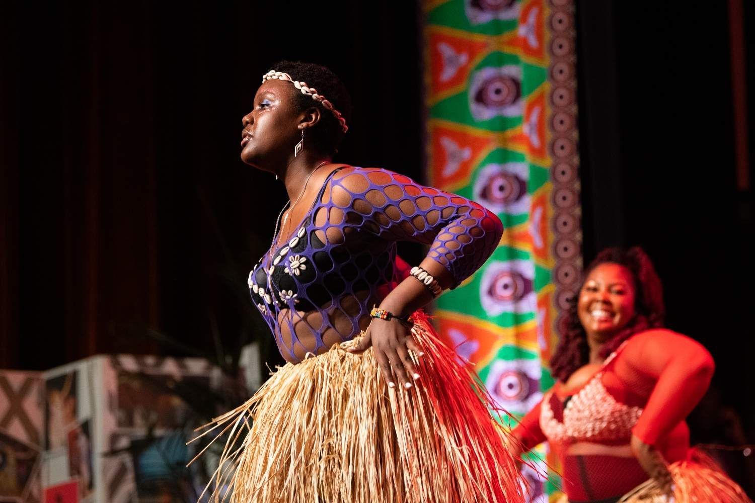 Two Black women dancers in traditional costumes onstage, one in blue, one in red, smiling.