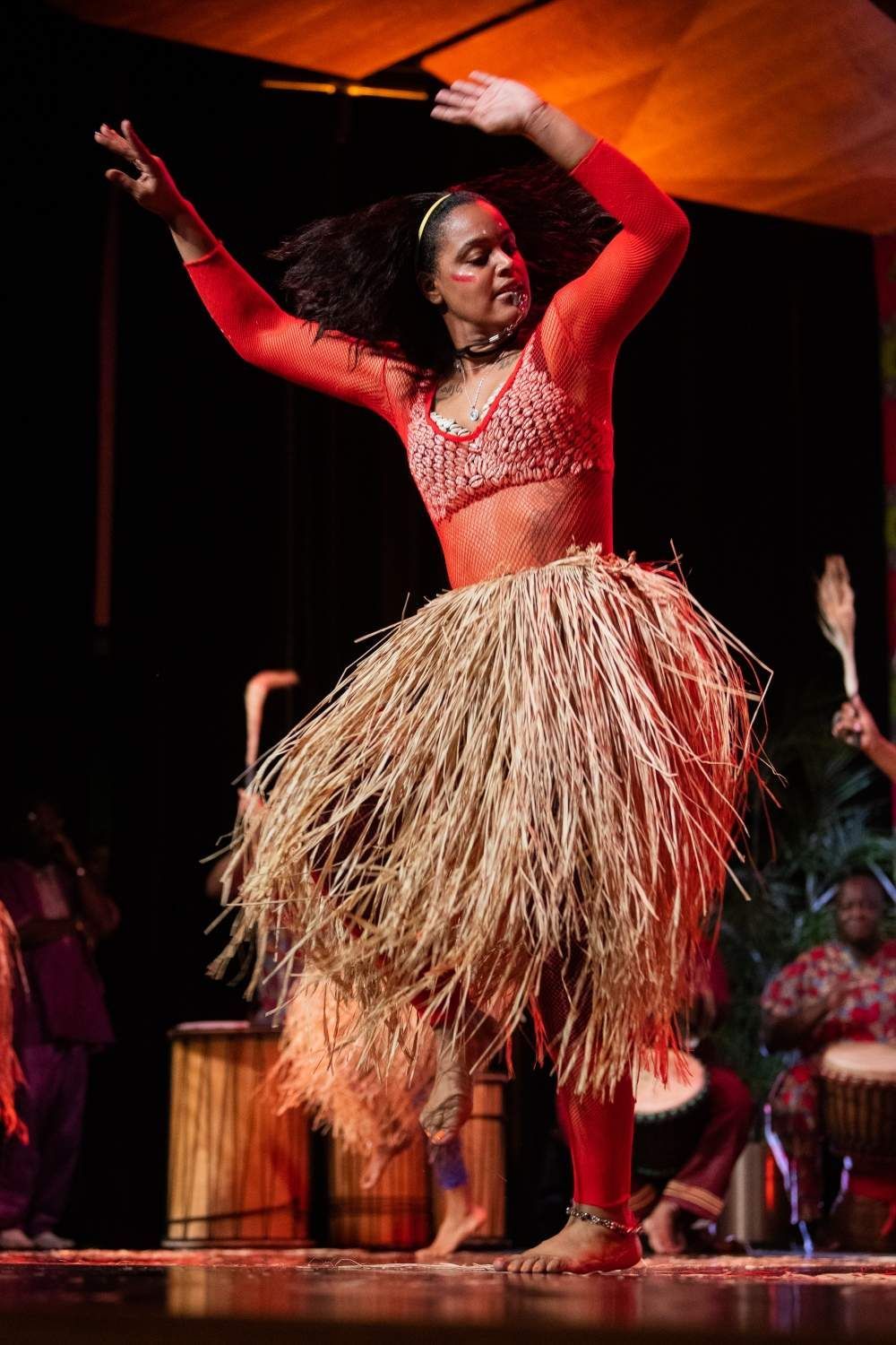 Woman in red outfit dances onstage, wearing a grass skirt.