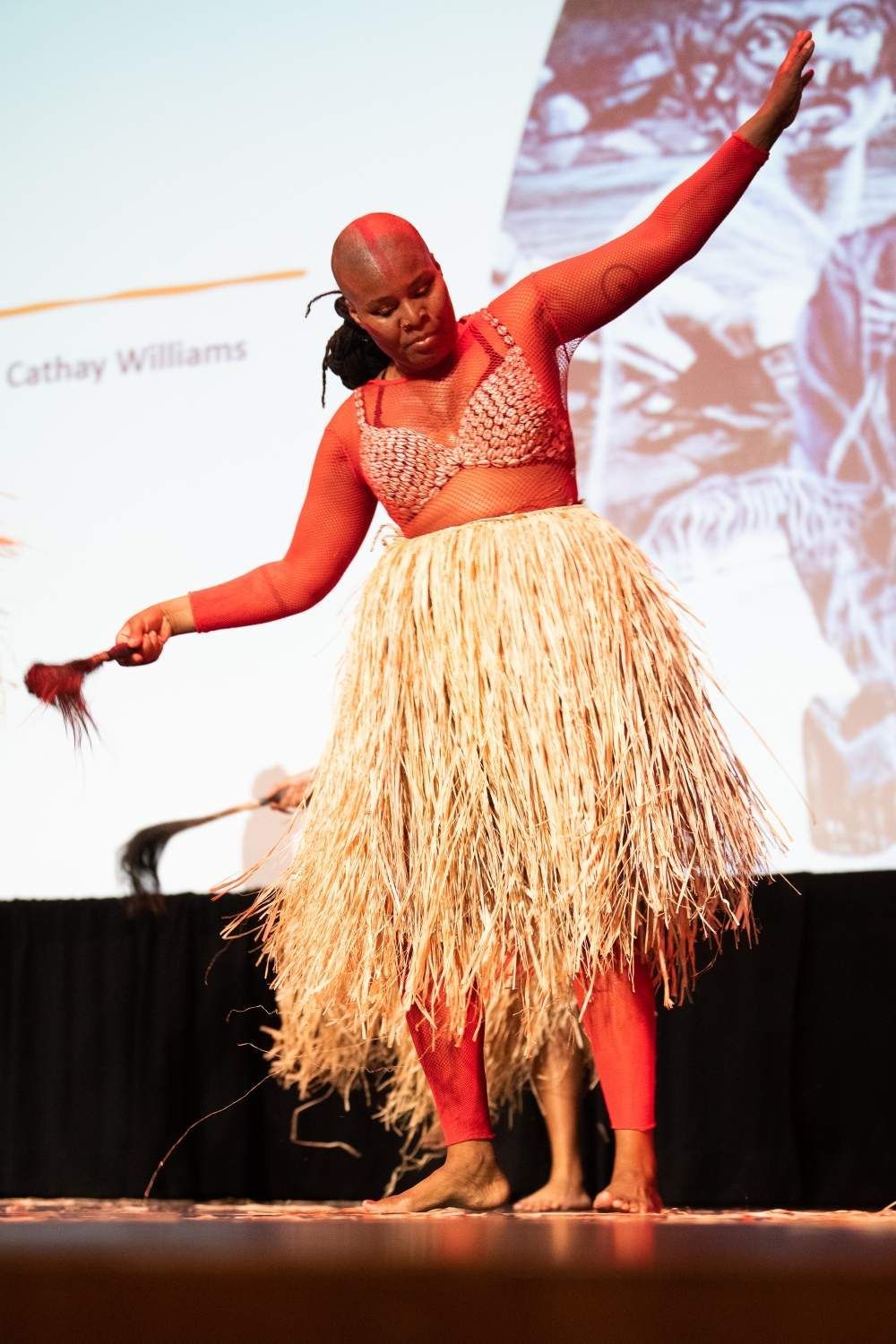 Woman in red, dancing on stage, wearing a straw skirt, holding a fan.