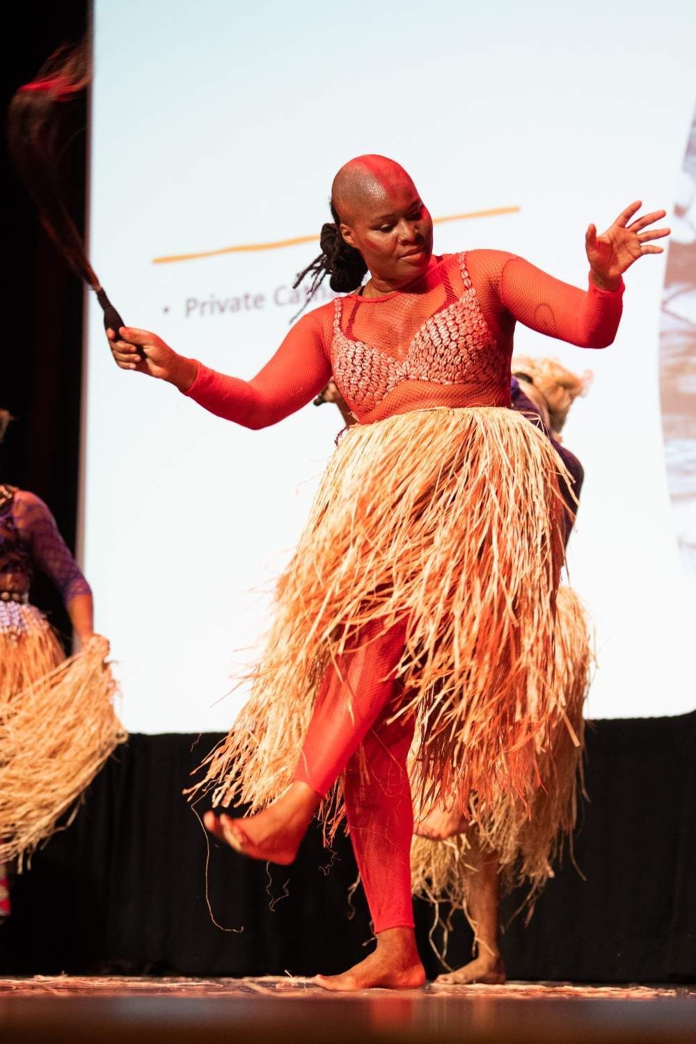 Woman in red costume and straw skirt dances, holding whip, onstage.