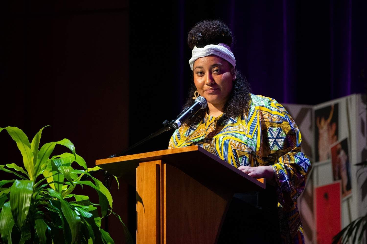Woman speaking at a podium, wearing colorful patterned clothing, with a plant to the left.