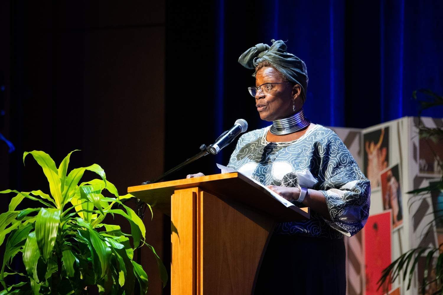 Woman in patterned attire speaking at a podium, on stage, with plant and decorated backdrop.