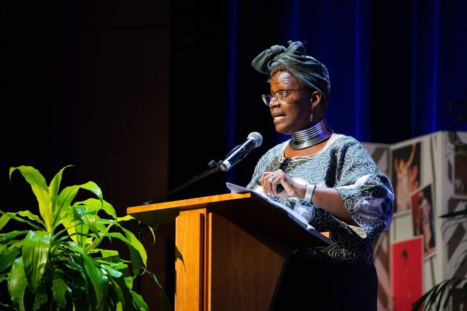 Woman speaking at a podium, wearing patterned headwrap and top, in a theater.
