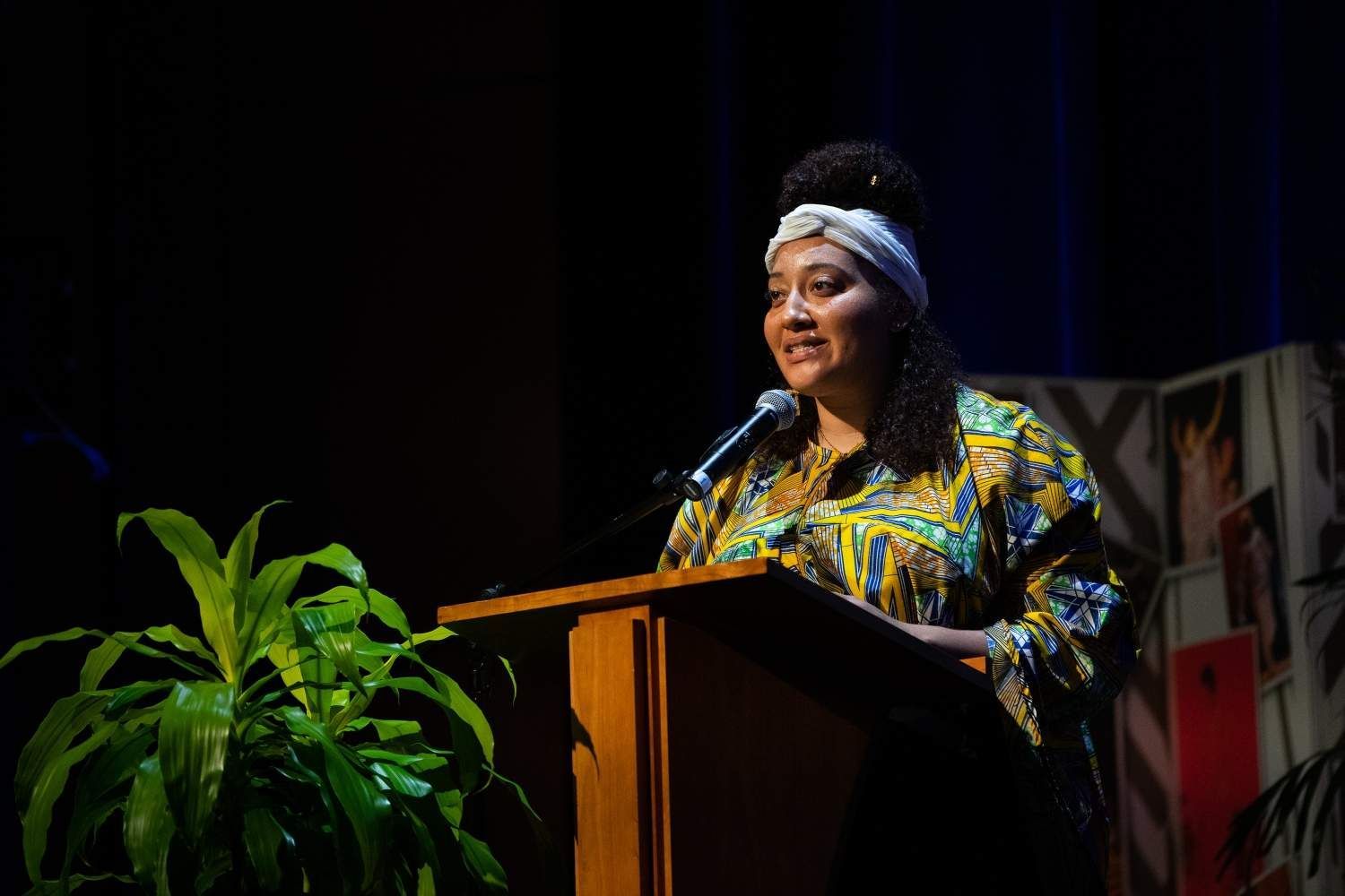 Woman speaking at a podium, wearing patterned shirt and headband. Stage lit, plants on left.