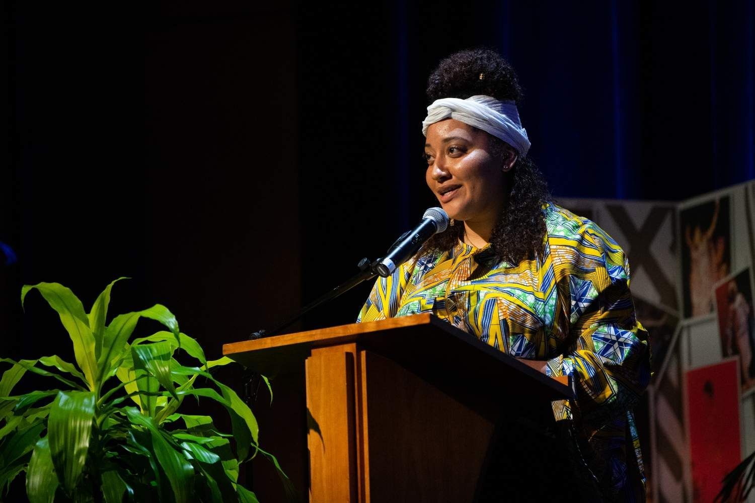 Woman in colorful dress speaks at a podium. She has a white headband and is in a dark auditorium.
