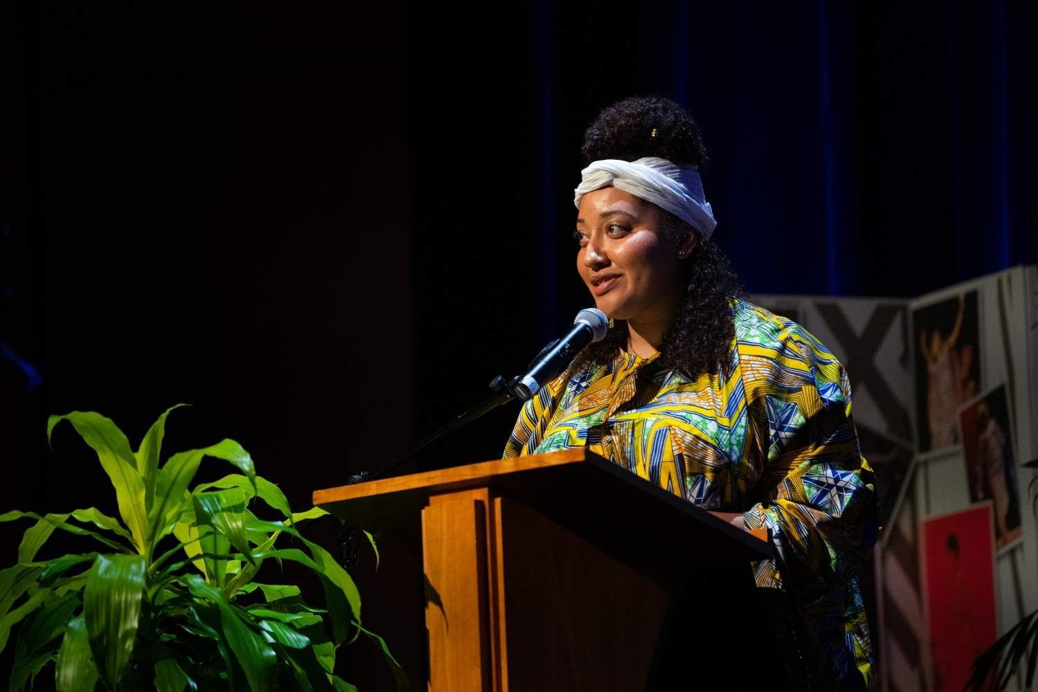 Woman speaking at a podium, wearing patterned shirt and headwrap. Dark stage, plant and decorations visible.