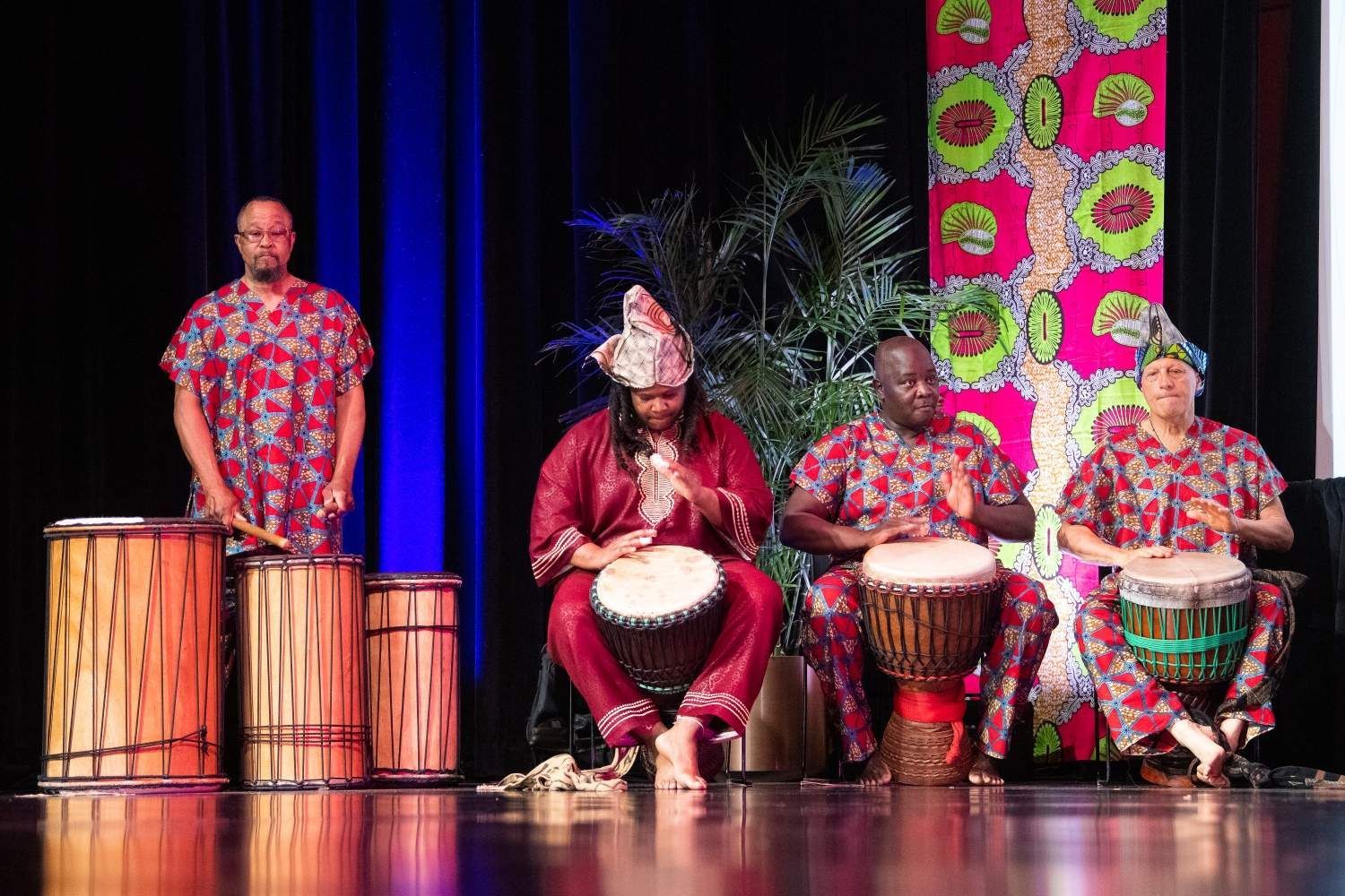 Four people in red patterned shirts playing drums on a stage, with a colorful fabric backdrop.