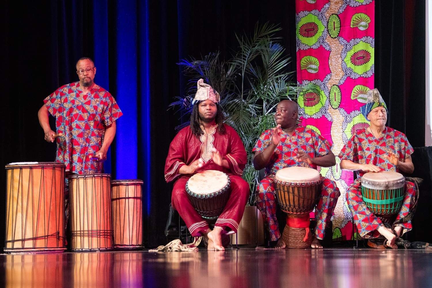 Four people on stage playing drums, wearing colorful African print outfits.