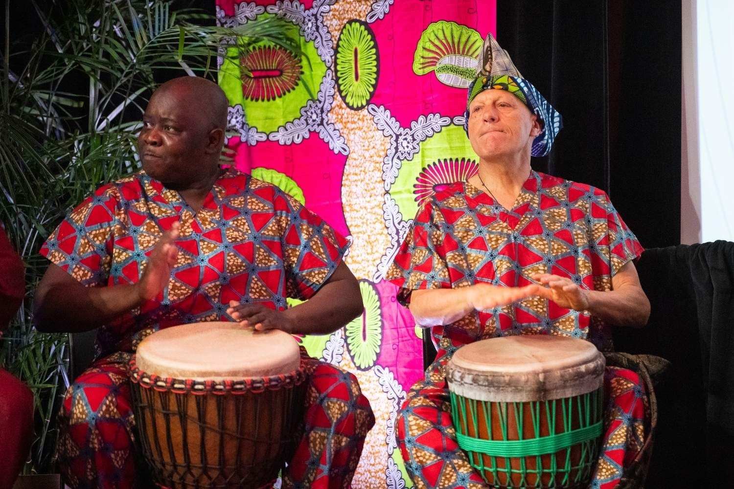 Two people in matching African print outfits play drums, smiling, with colorful fabric backdrop.