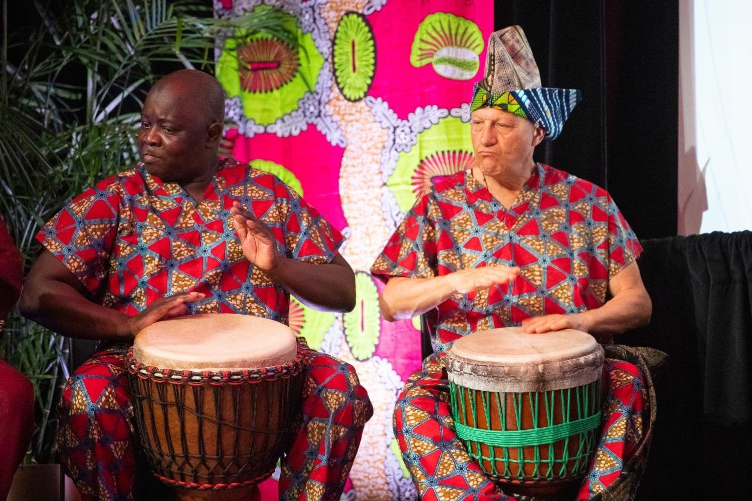 Two men in African attire play drums in front of colorful fabric.