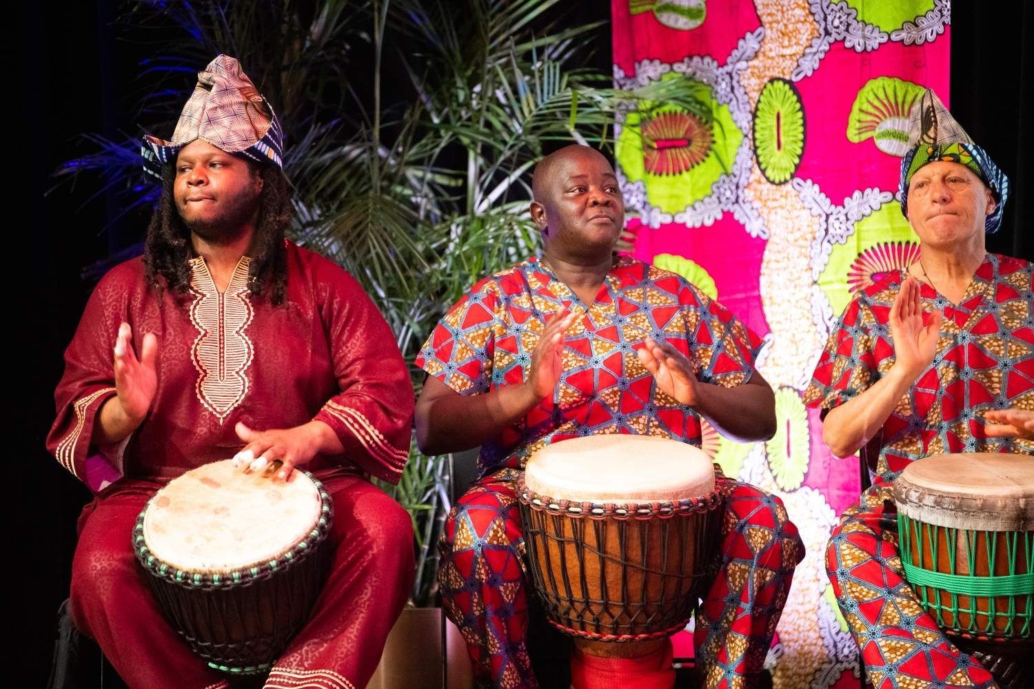 Three people playing drums, wearing colorful African clothing; vibrant stage backdrop.
