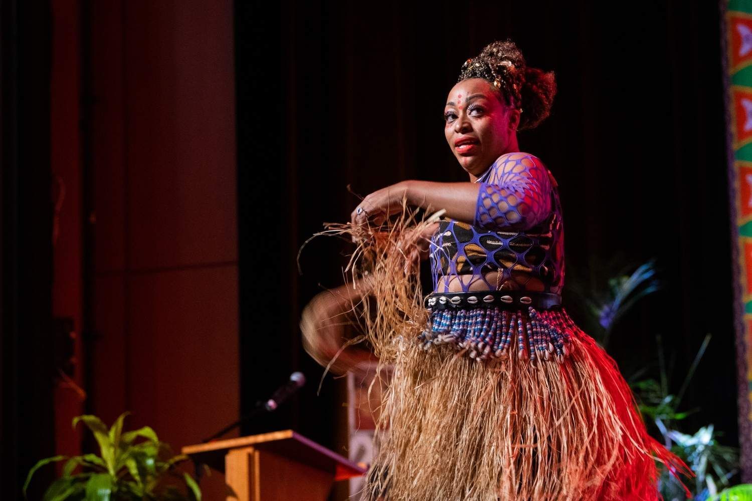 Woman in colorful African garb dances on stage, holding a gourd.