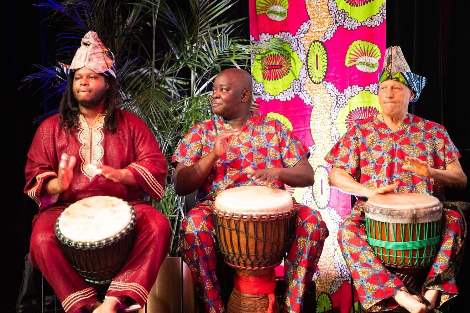 Three people in African attire play drums onstage; colorful backdrop.