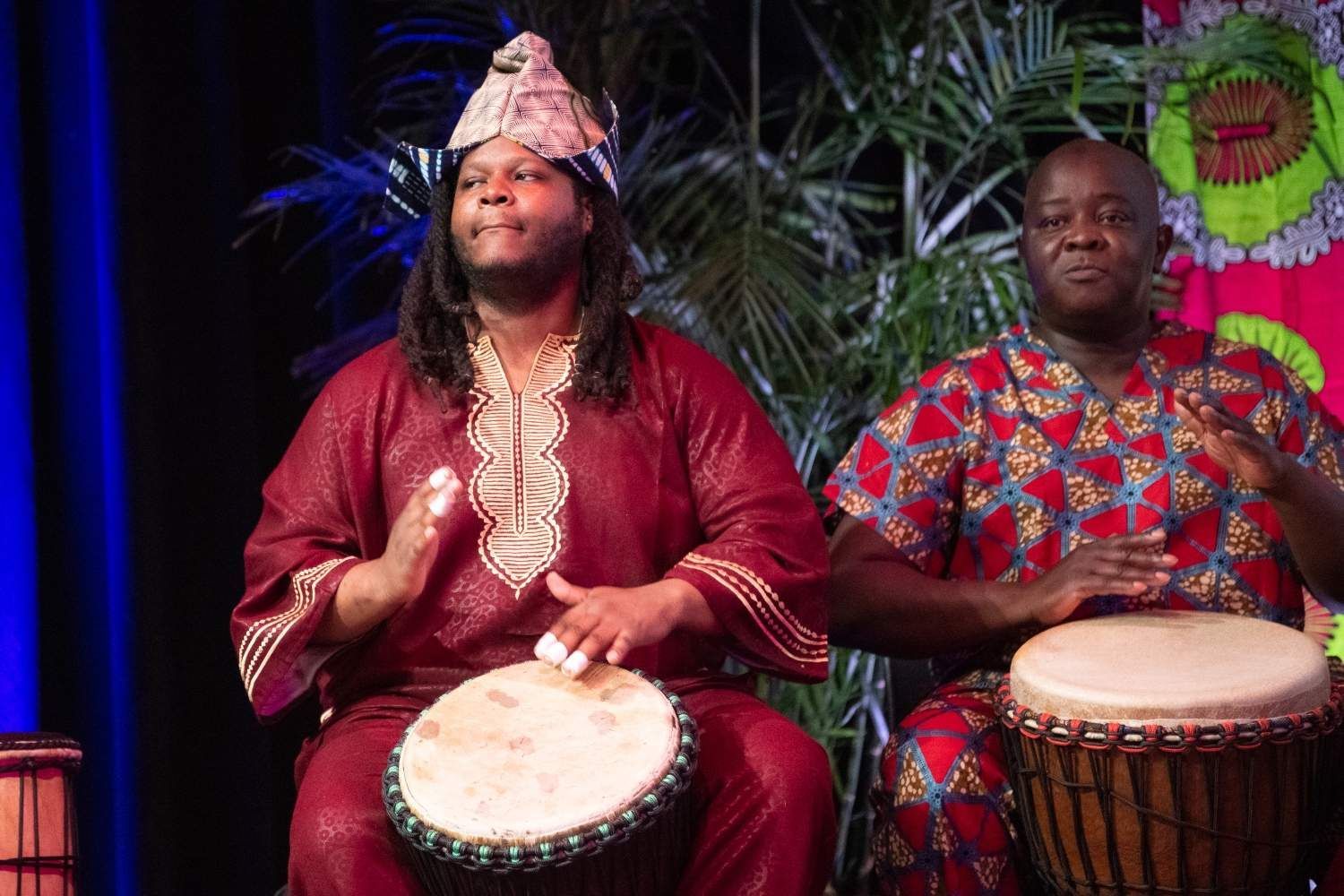 Two men in colorful African attire playing drums on stage.