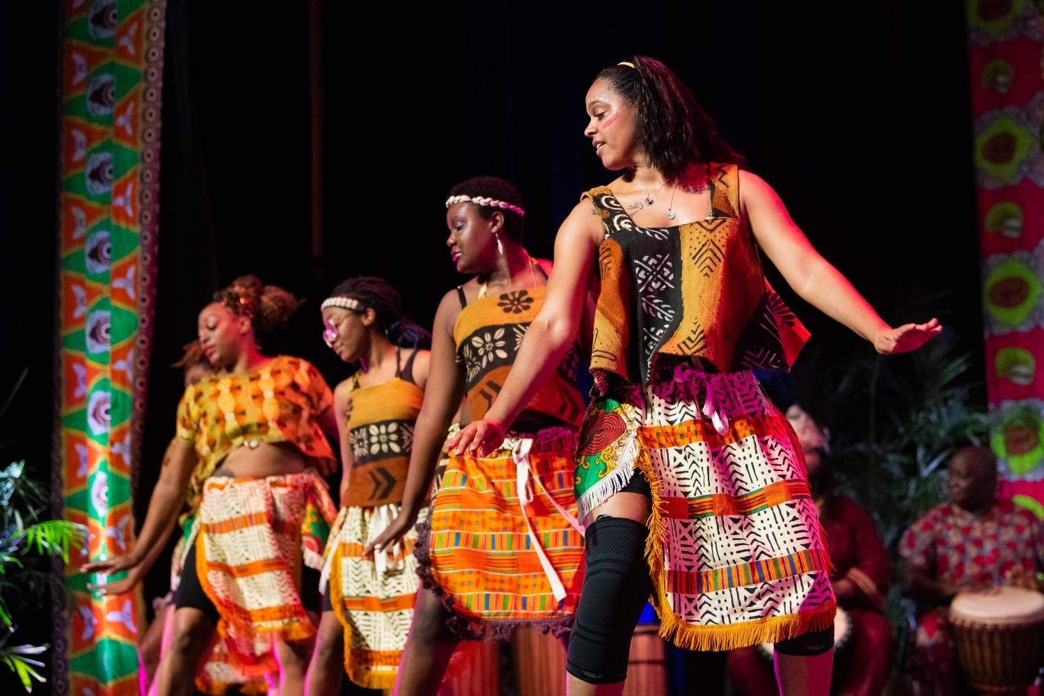 Dancers in colorful African attire on stage, performing a dance with joy; drummer in the background.