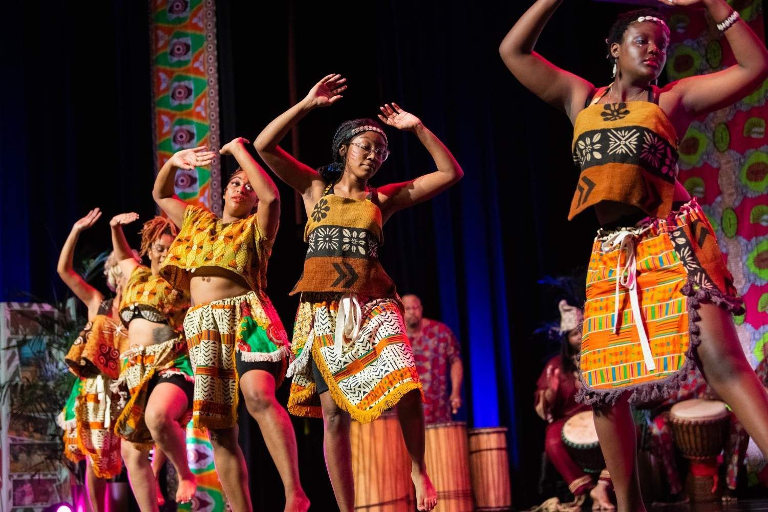 African dancers in colorful outfits, performing on stage with drummers in the background.