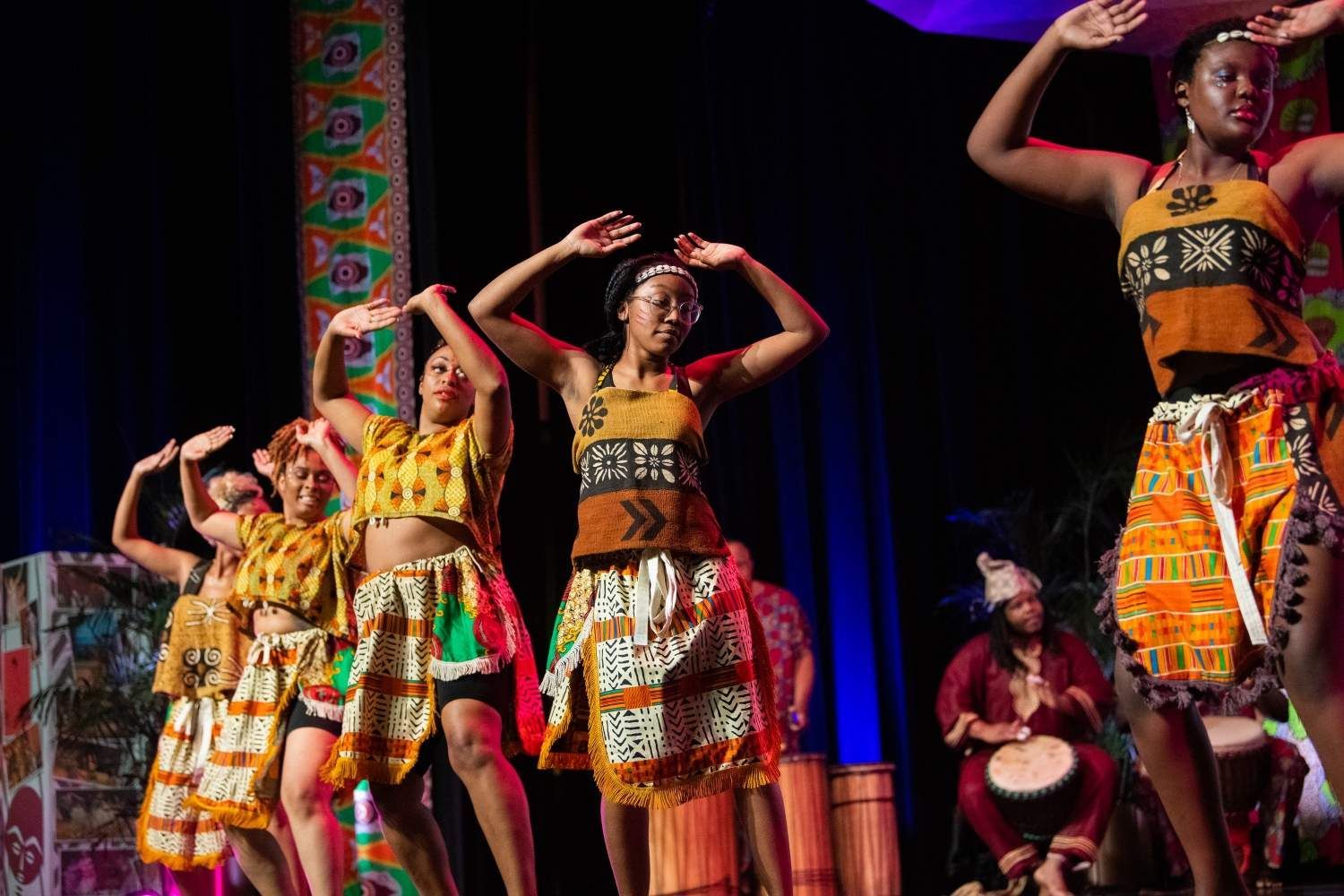 Dancers in colorful African attire perform onstage with a drummer; arms raised.