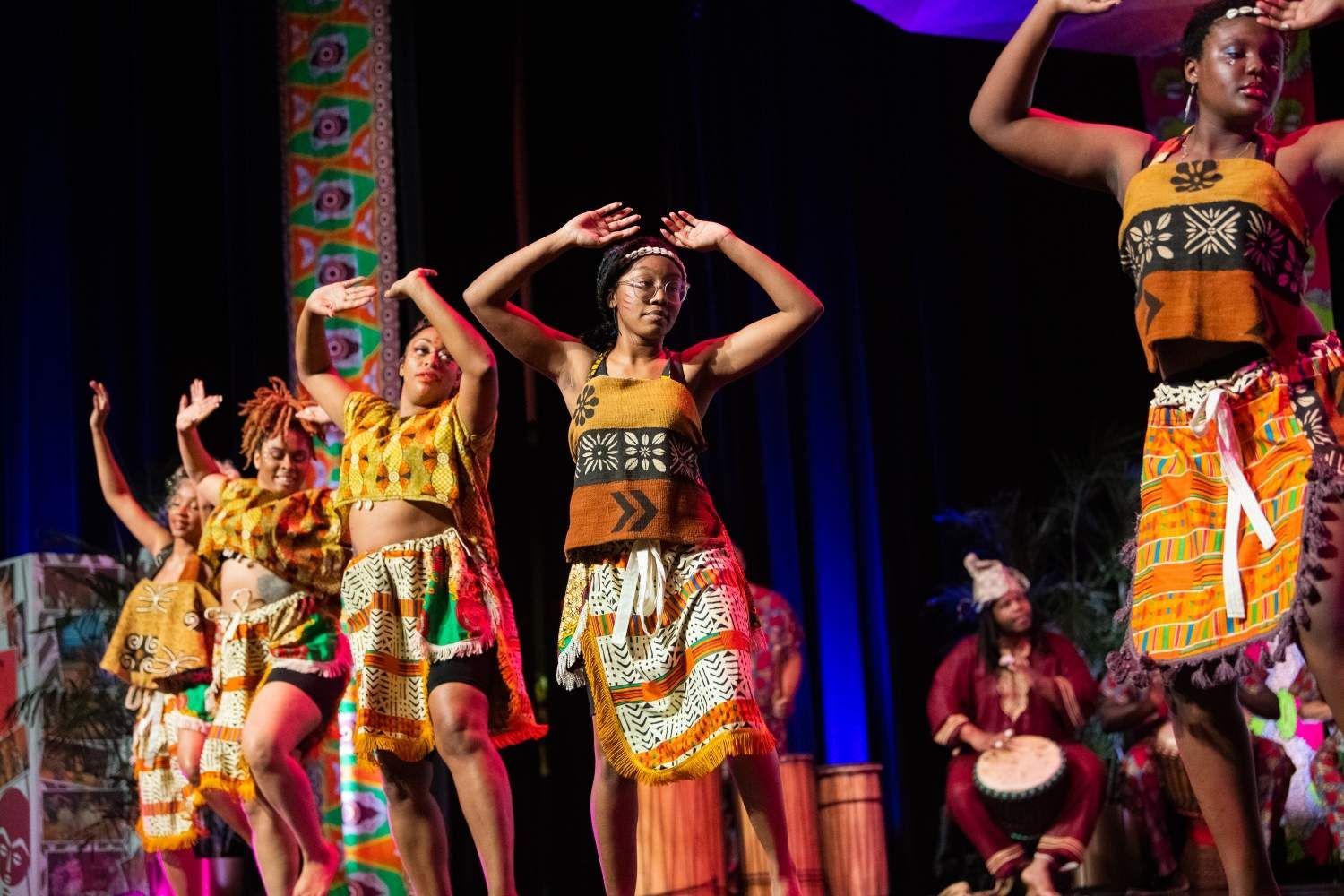 Dancers in colorful African attire perform on stage with a drummer, arms raised.