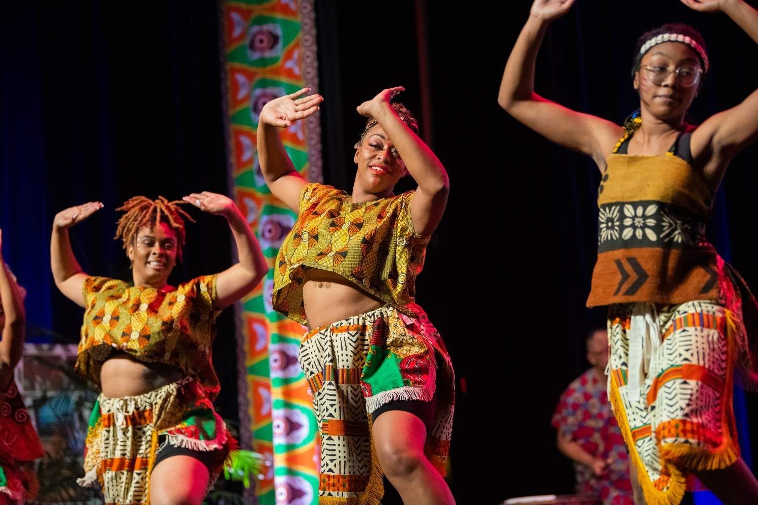 Three Black women dancing onstage in colorful African attire with arms raised.