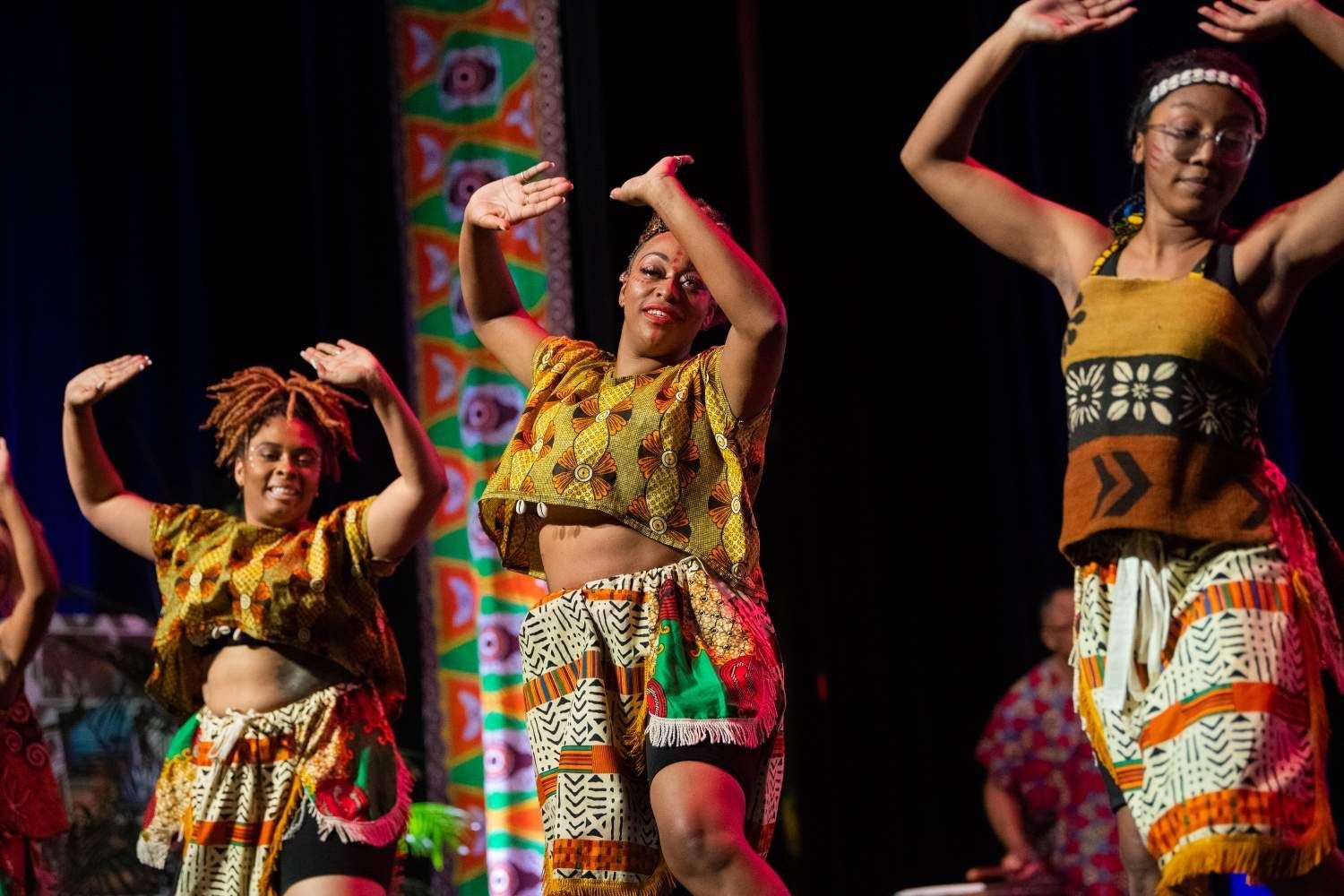 Dancers in vibrant African-print outfits perform on stage, arms raised.