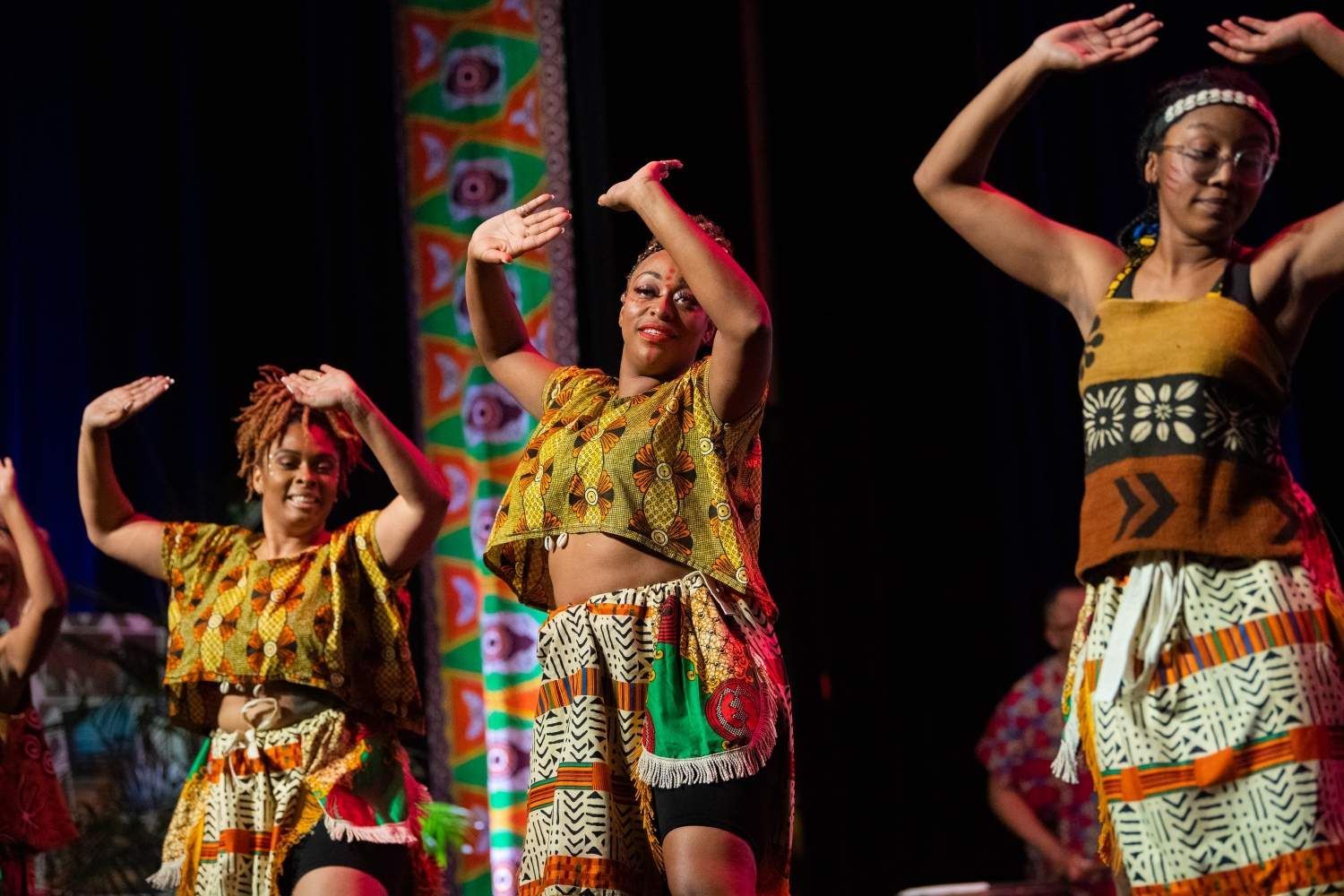 African dancers on stage, performing with raised arms, wearing colorful patterned outfits, dark background.