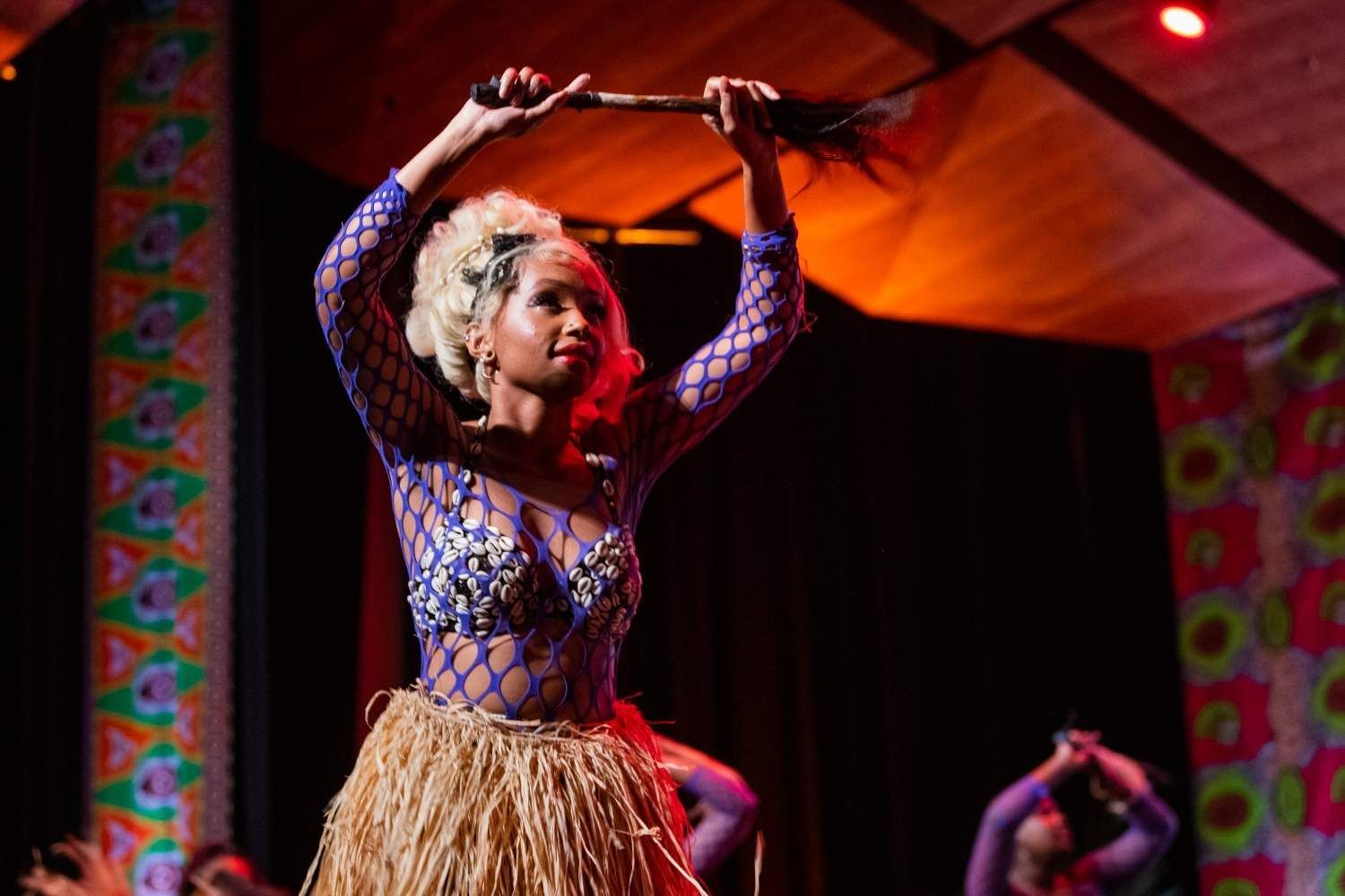 Woman in blue top and grass skirt performs dance, holding a stick overhead.