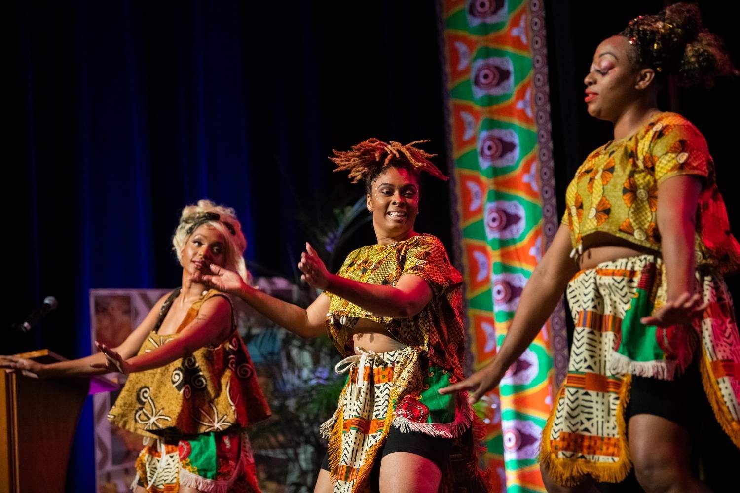 Three Black women dancing on stage, wearing colorful African print outfits.