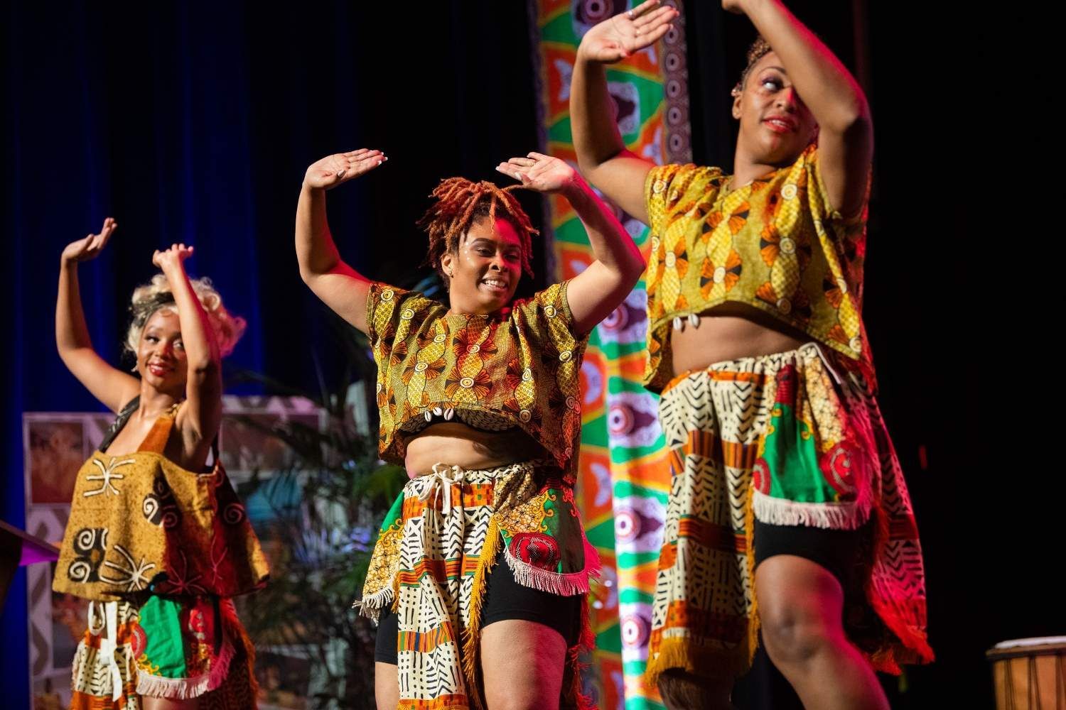 Three Black women dance on stage wearing colorful African-inspired clothing, arms raised, smiling.
