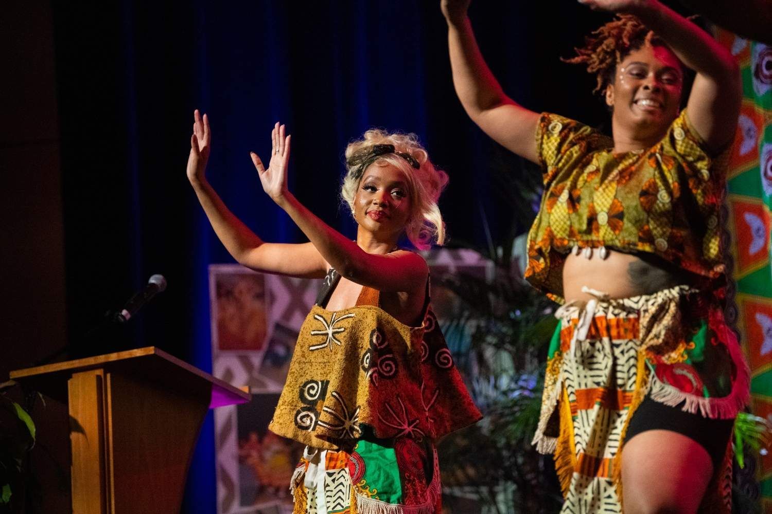 Two women dancing on stage in colorful African-inspired clothing, arms raised.