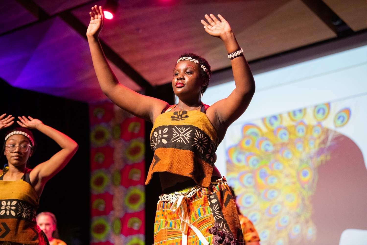 Woman in orange African attire dancing onstage, arms raised.