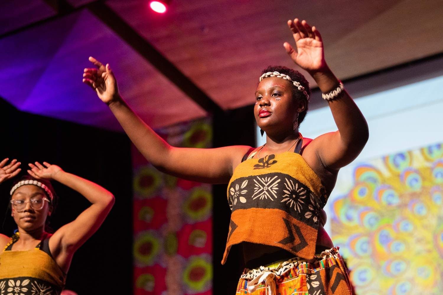 Two women performing an African dance on stage, arms raised, wearing patterned dresses, headbands, and bracelets.