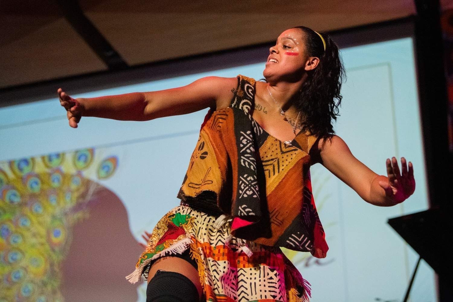 Woman dancing on stage with arms outstretched, colorful patterned clothing, projected peacock.
