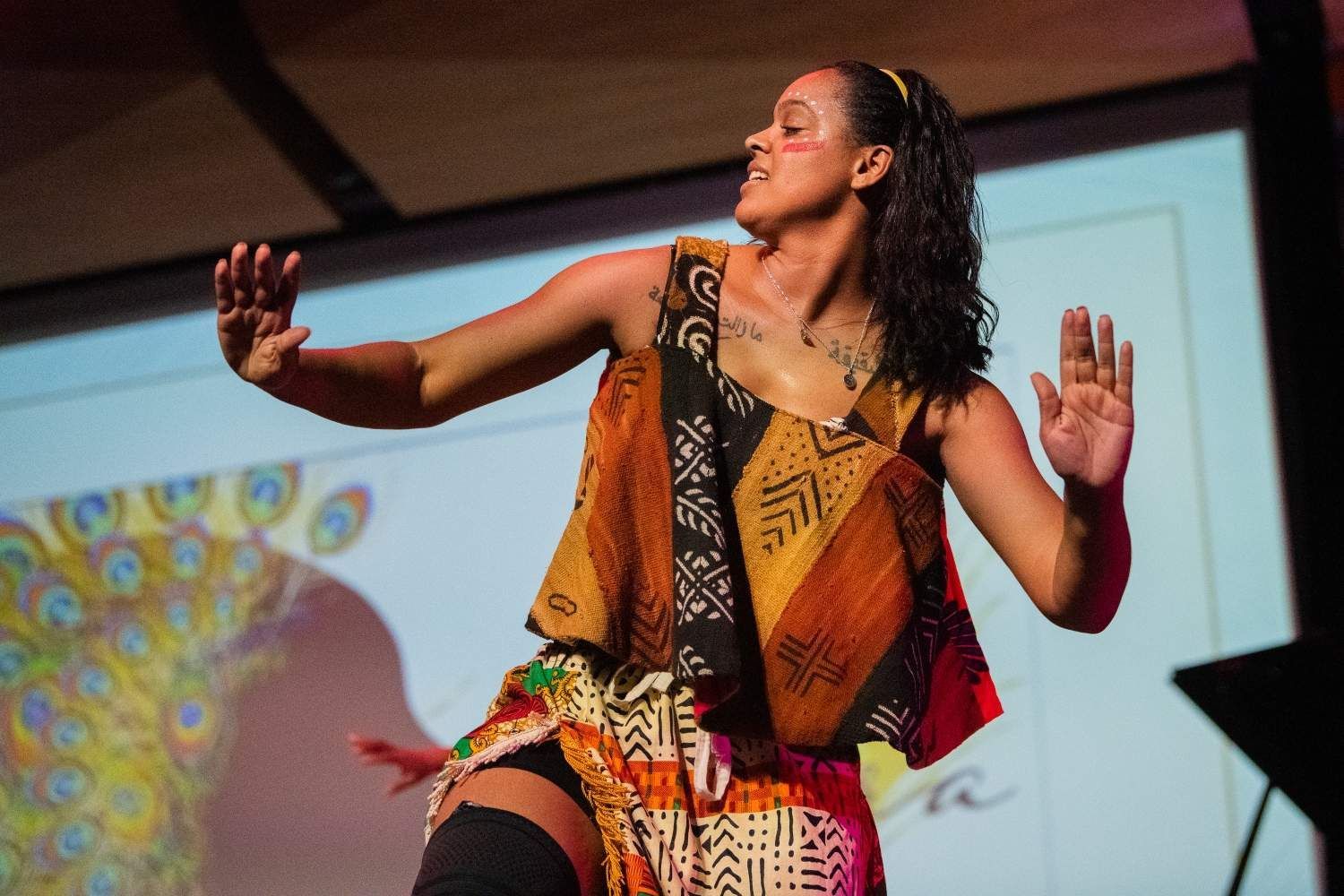 Woman performing a dance on a stage, arms outstretched. She wears colorful patterned clothing.
