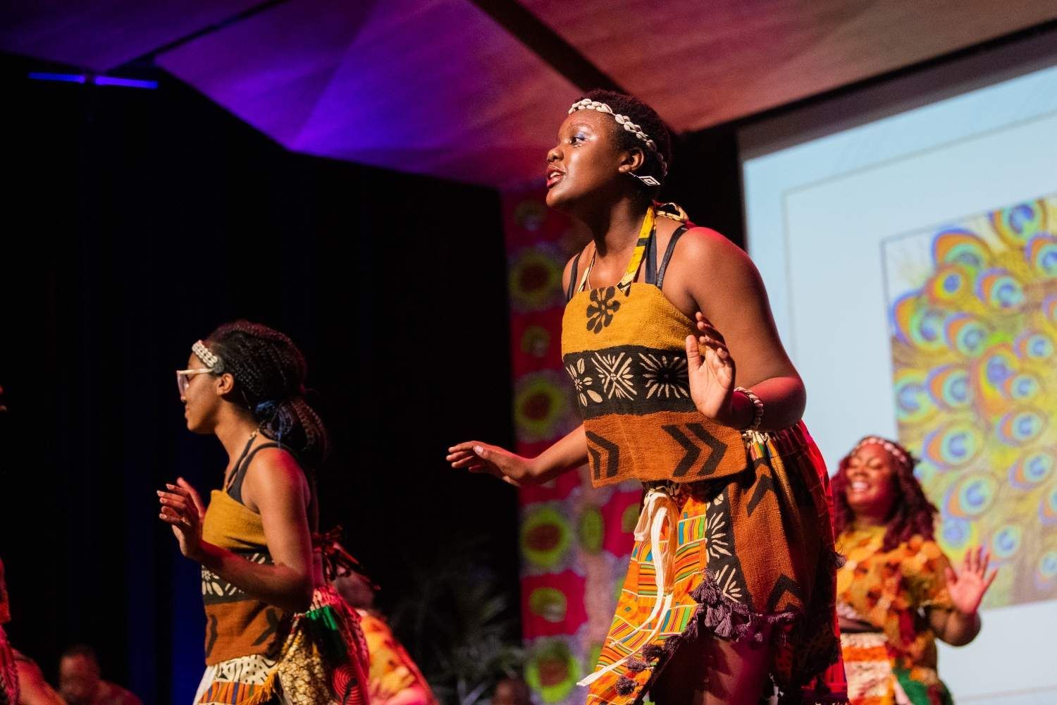 Three women in traditional African outfits sing and dance on stage, bright colors, floral background.