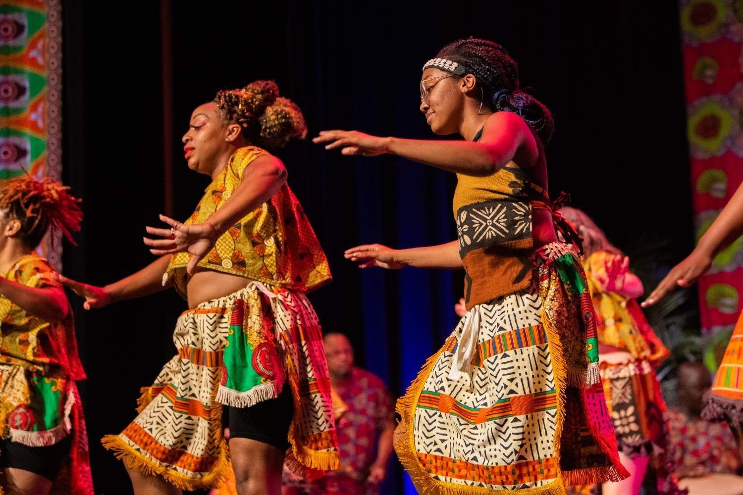 Dancers in colorful African attire perform on a stage with dynamic movements, lit by stage lights.