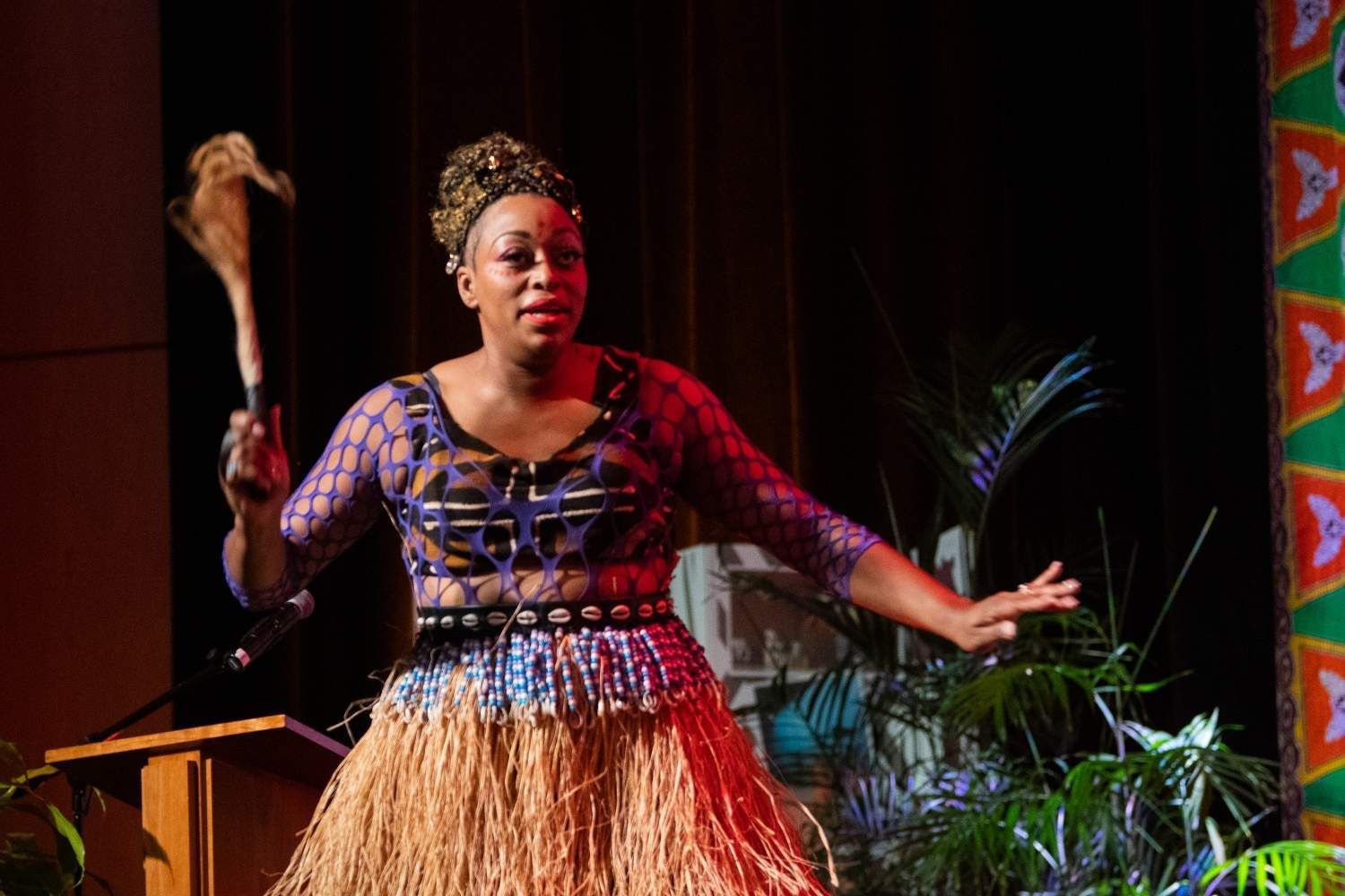 Woman in traditional dress holding a club on stage.  Dark blue, brown, and tan clothing.  Palm trees in the background.