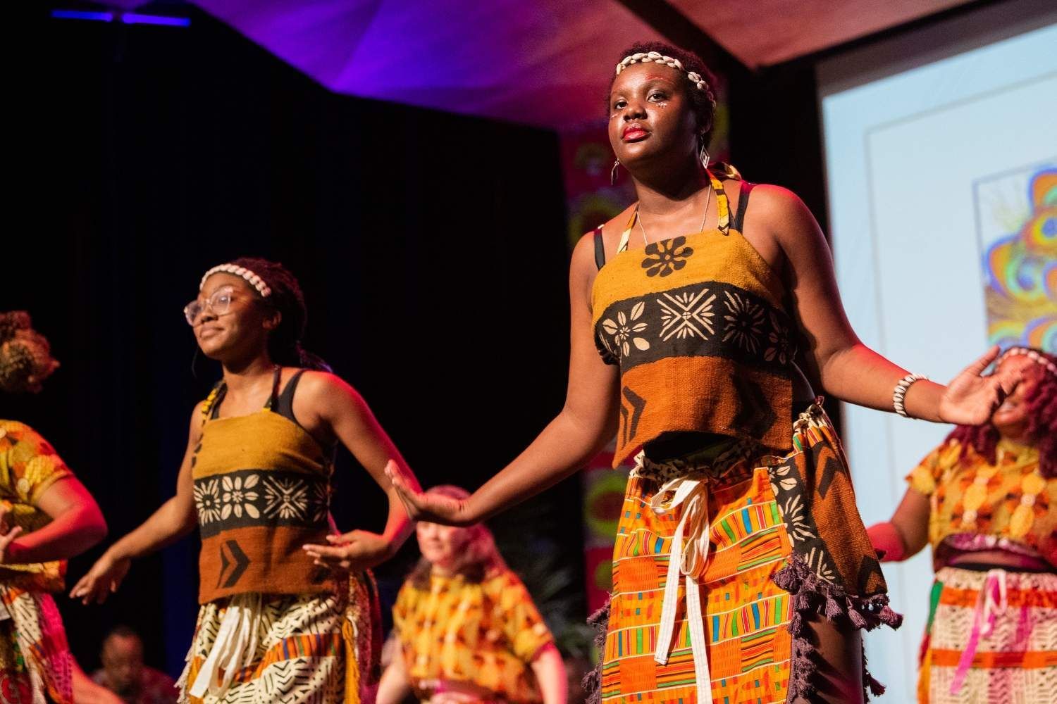 Women in traditional African outfits perform on stage, arms outstretched.