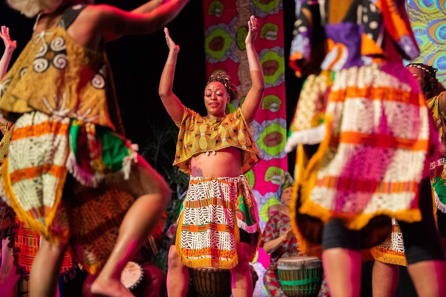 Dancers in colorful African-style attire, arms raised, performing on a stage with drums and vibrant decorations.