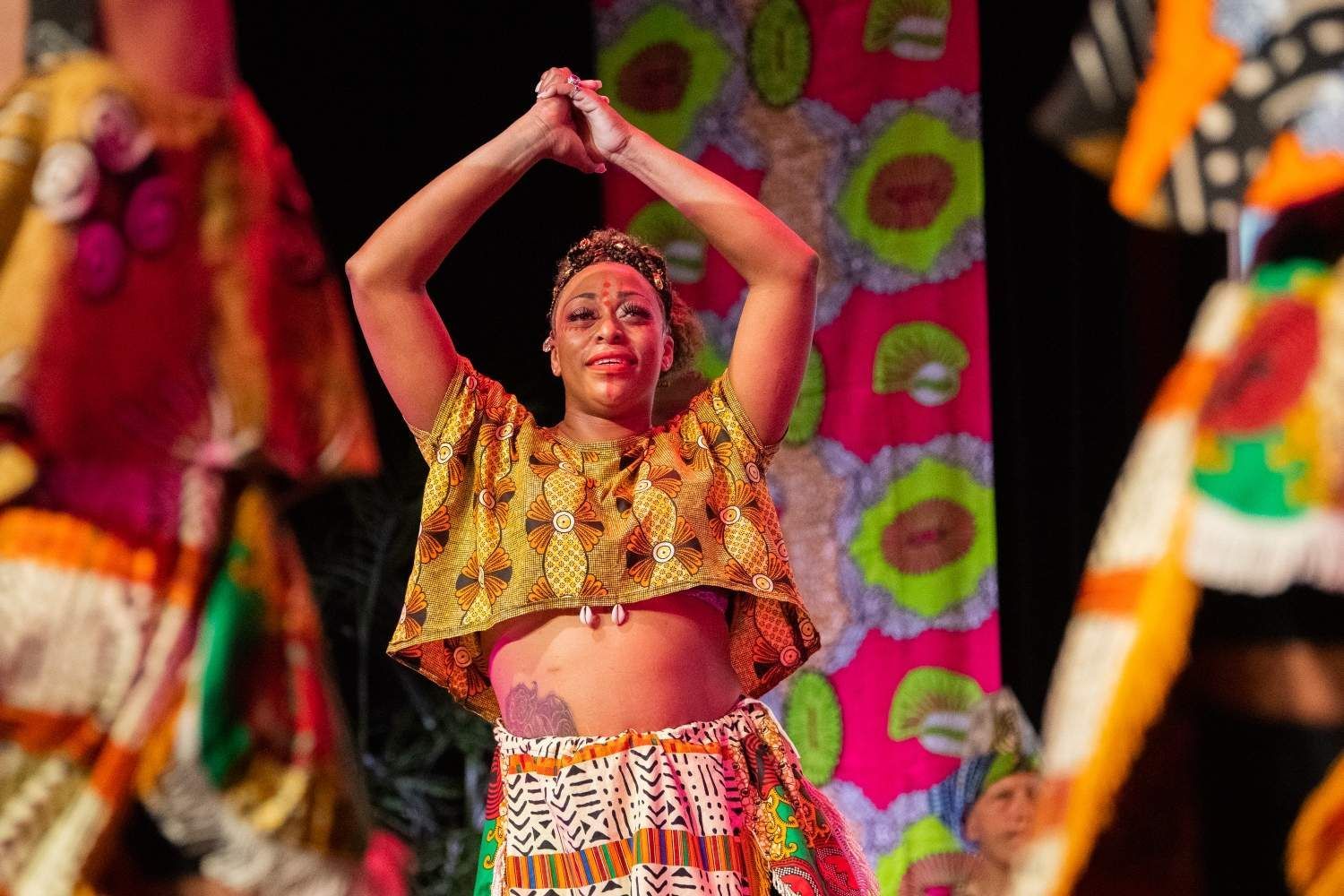 Woman in yellow patterned top and skirt dances on stage, arms raised.