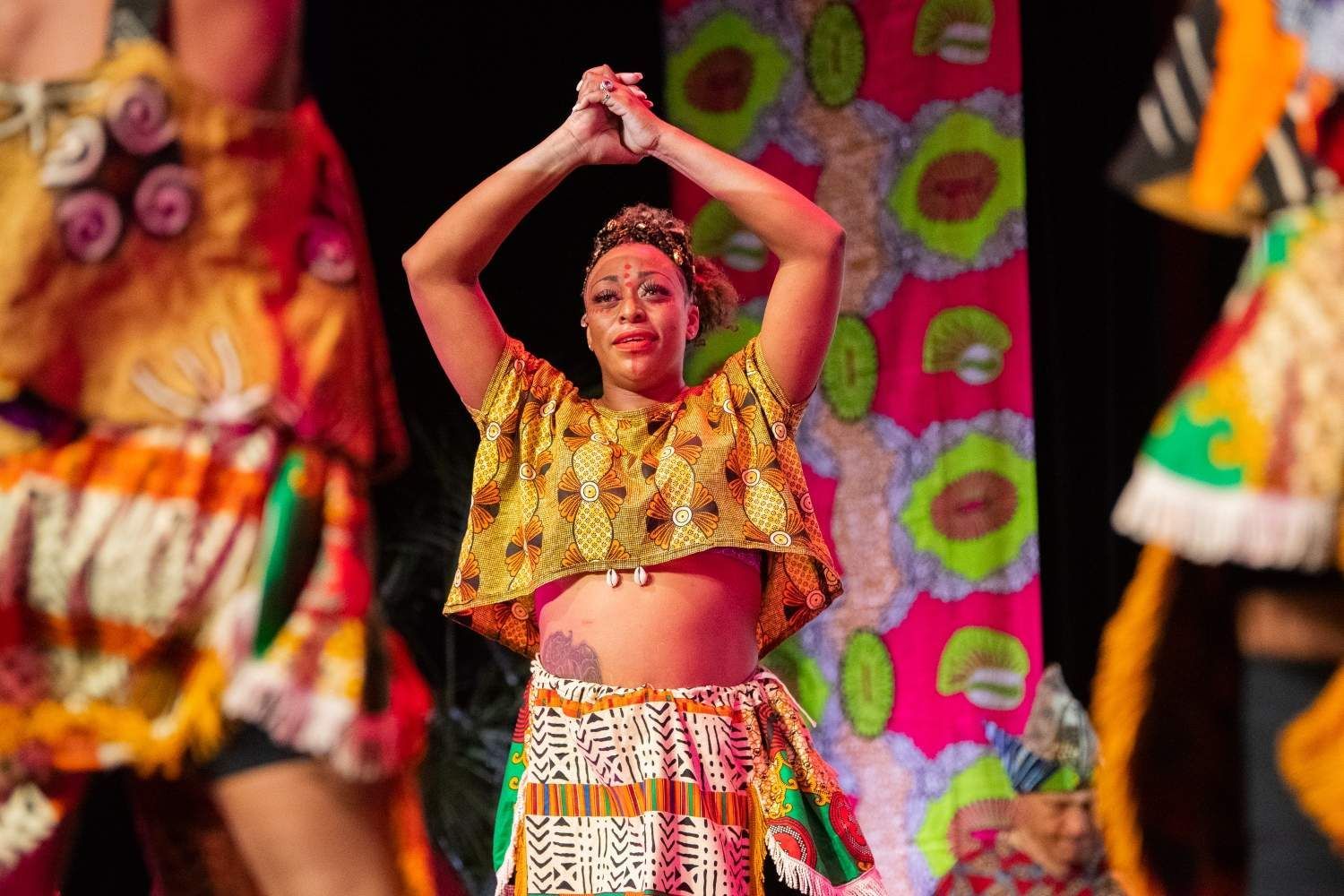 Woman in African-print outfit dancing with arms raised, a colorful stage backdrop.