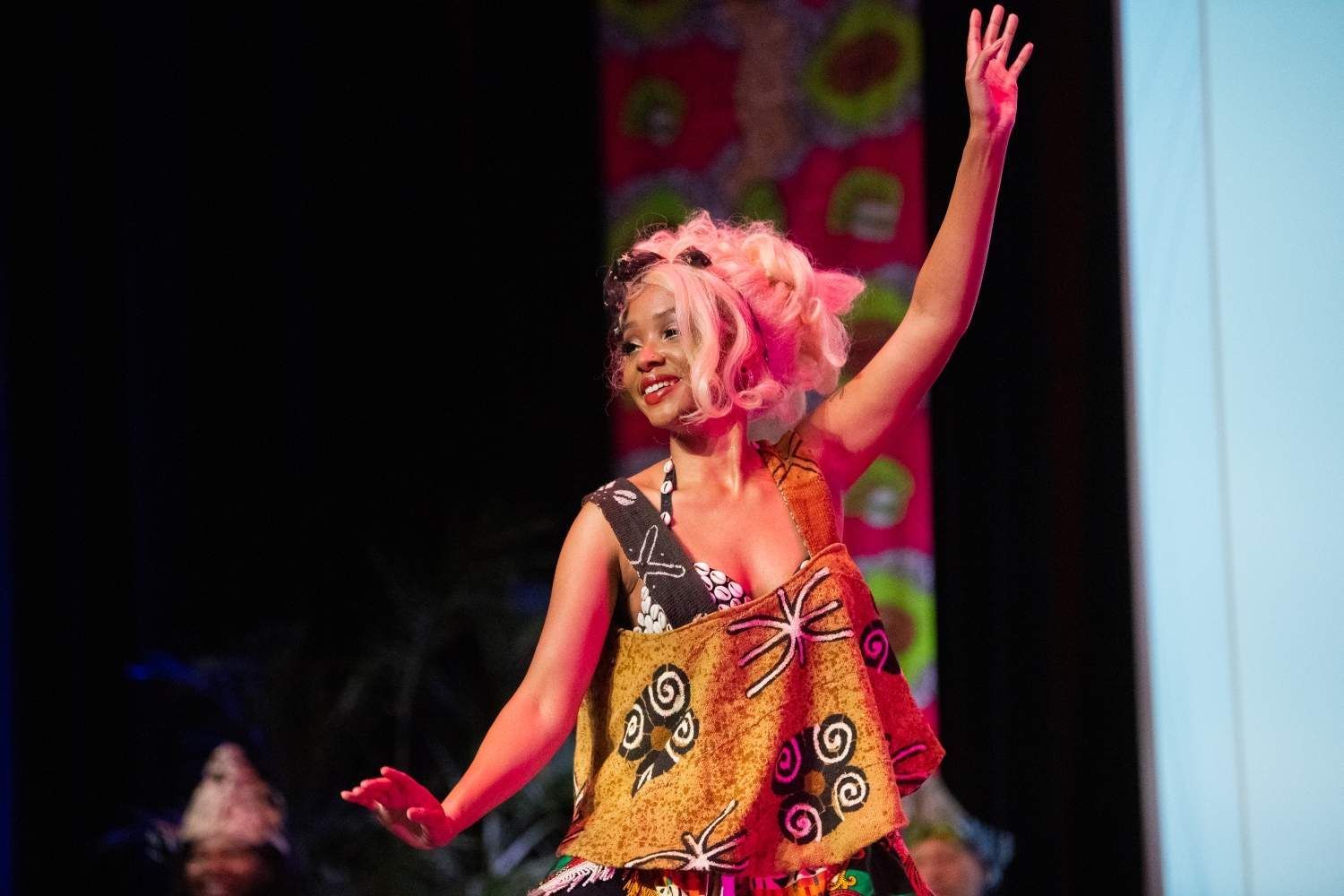 Woman in African print dress with pink hair, dancing on stage with her arm raised.