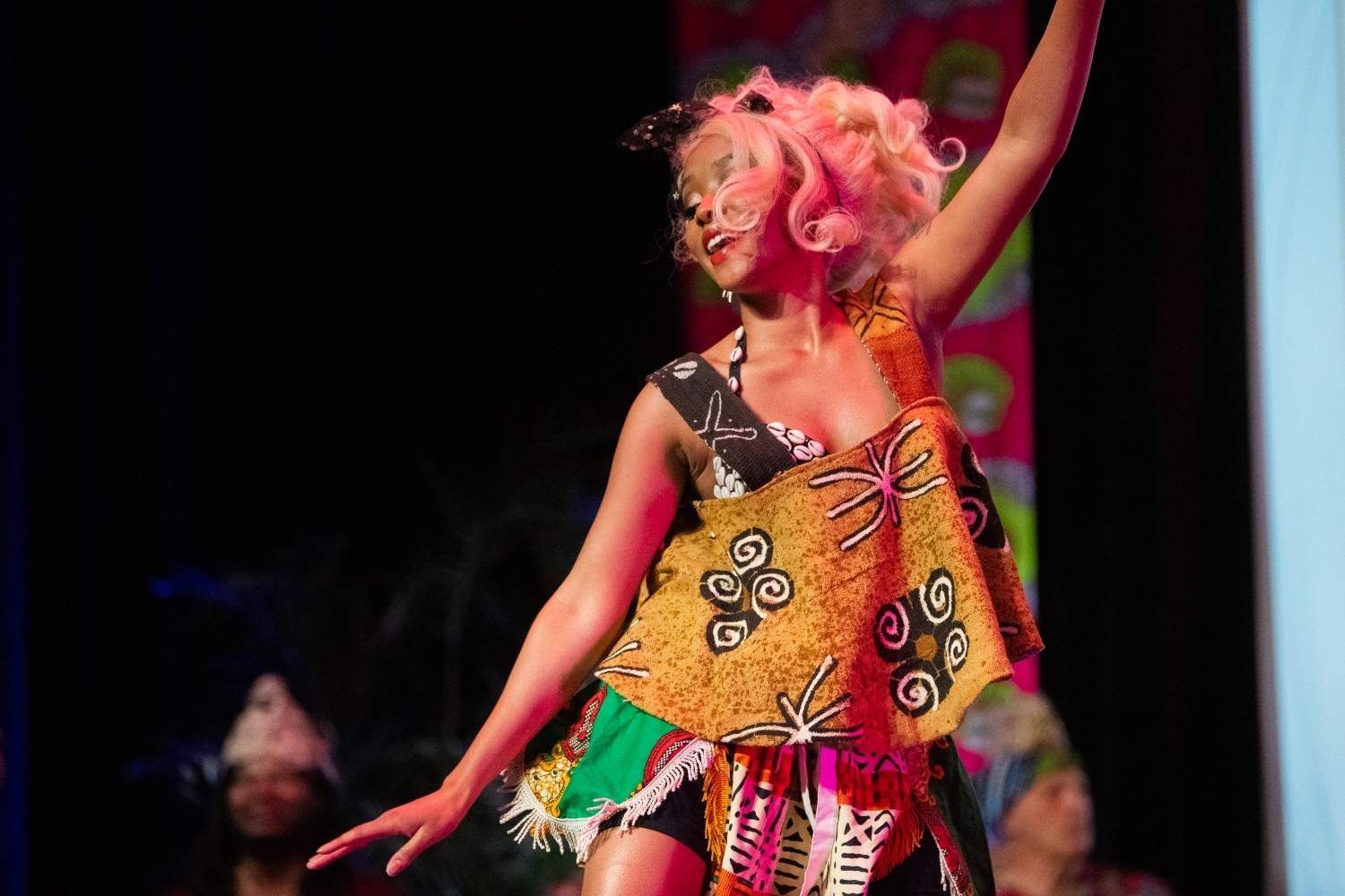Woman in colorful African-print outfit on stage, arms raised, smiling. Pink hair, black cat ears.