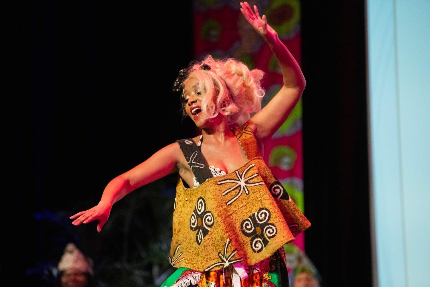 Woman in colorful African-print outfit dances onstage, arms raised, with pink hair.