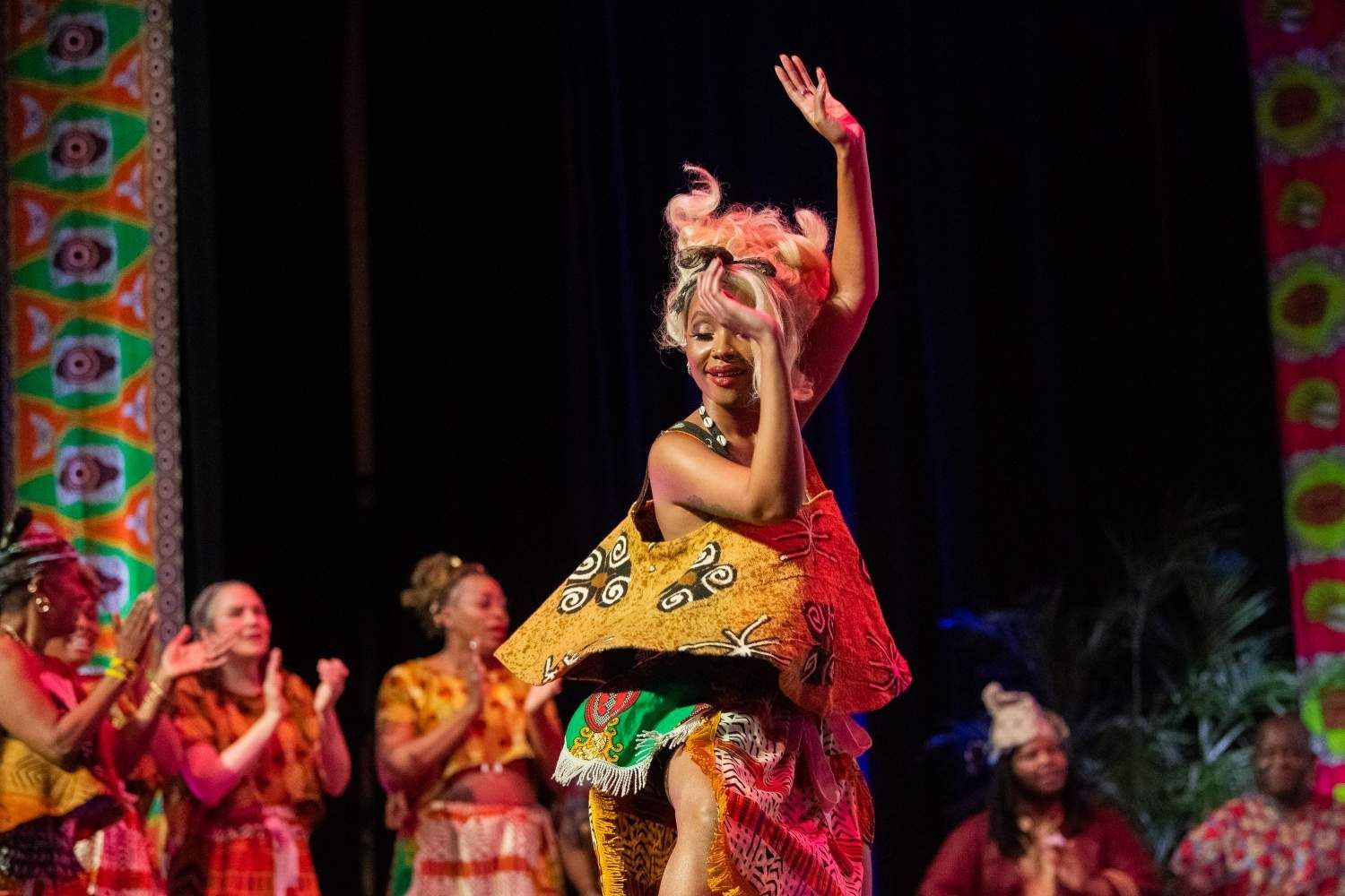 Woman in vibrant costume dances on stage; others clap, colorful backdrop.