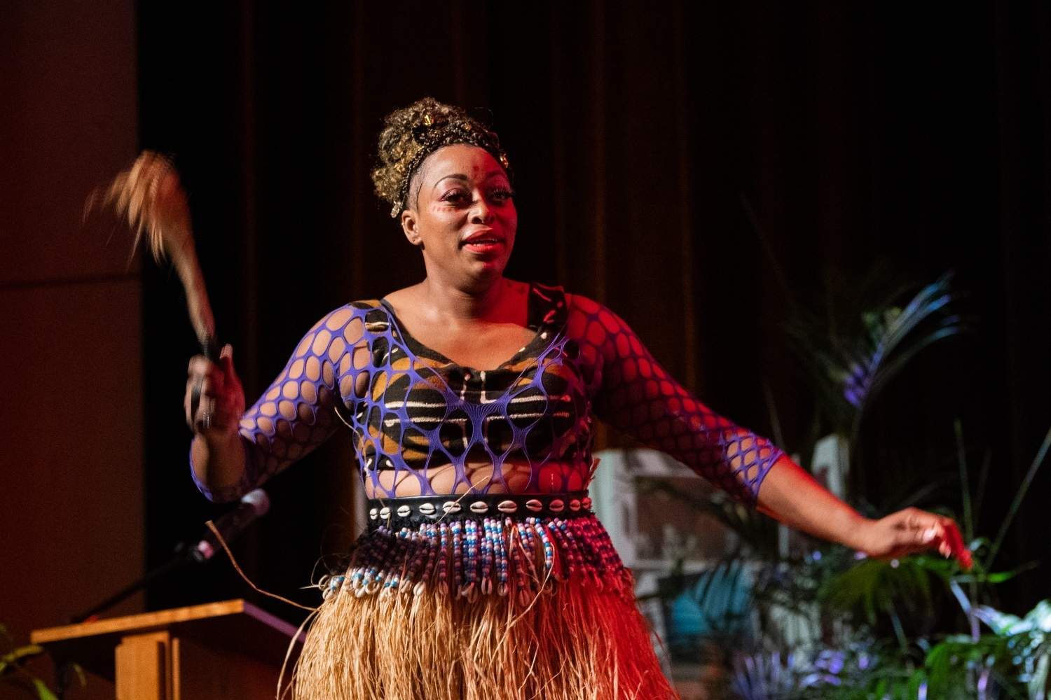 Woman in patterned dress and straw skirt holds a wand, performing onstage.