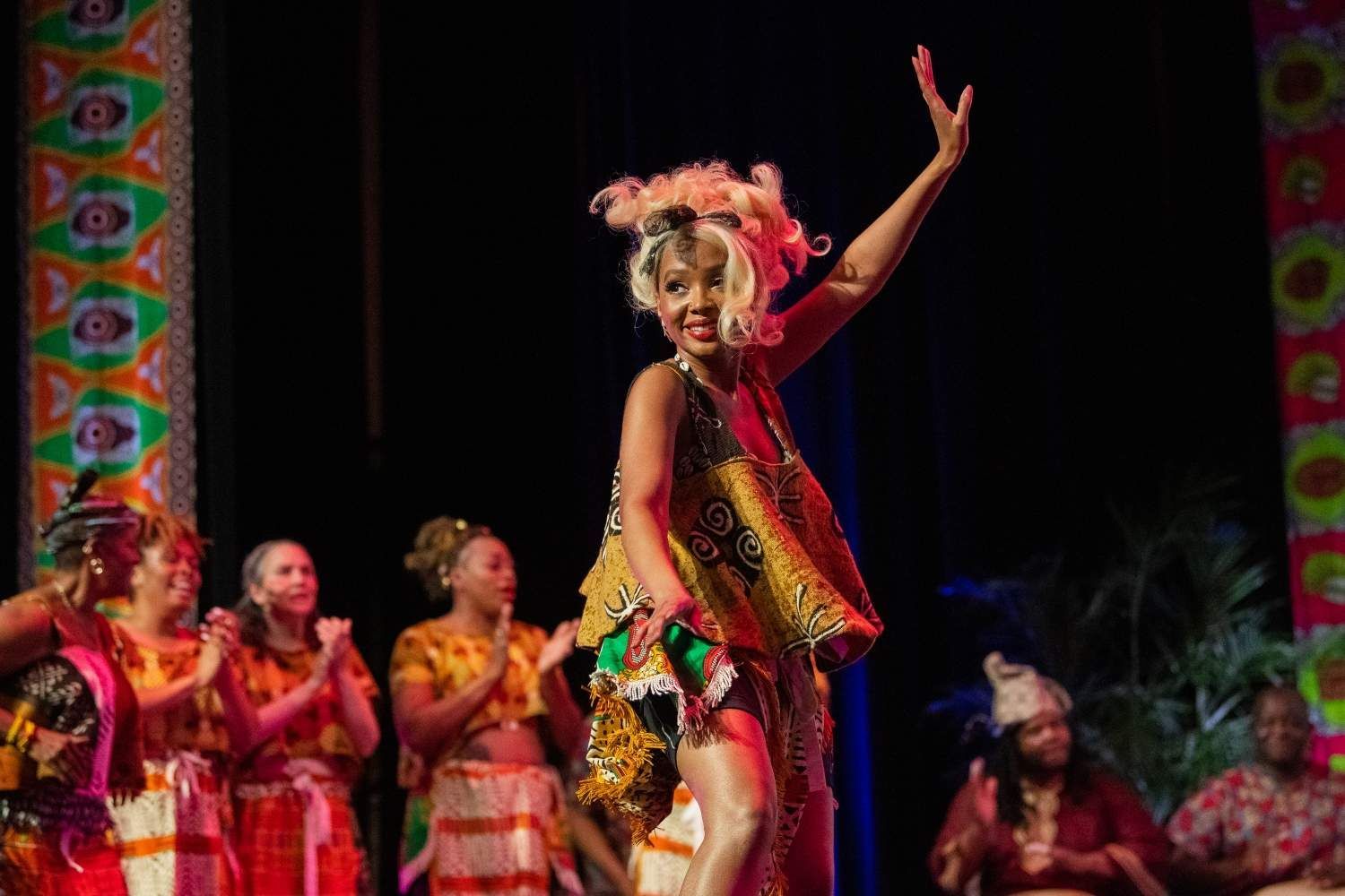 Woman in colorful dress dances on stage; other women clap.
