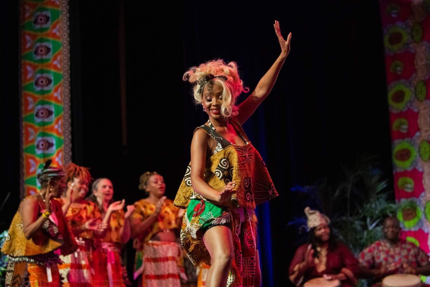 A Black dancer in colorful attire performs on stage with a band applauding.