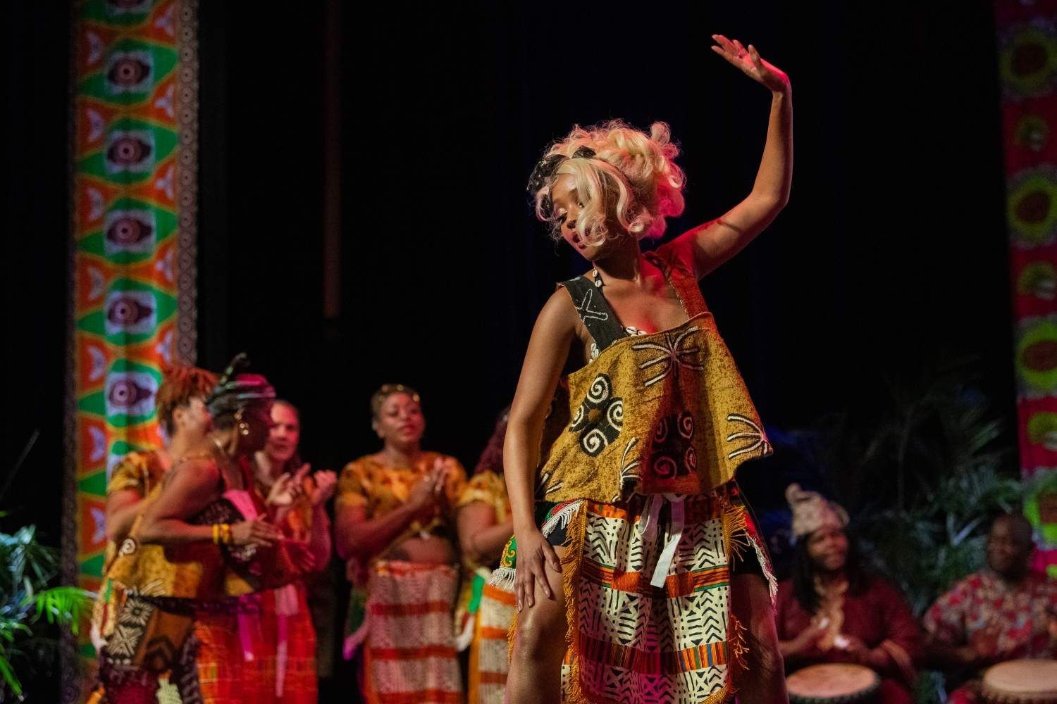 Woman in African print dress dances on stage, audience clapping.